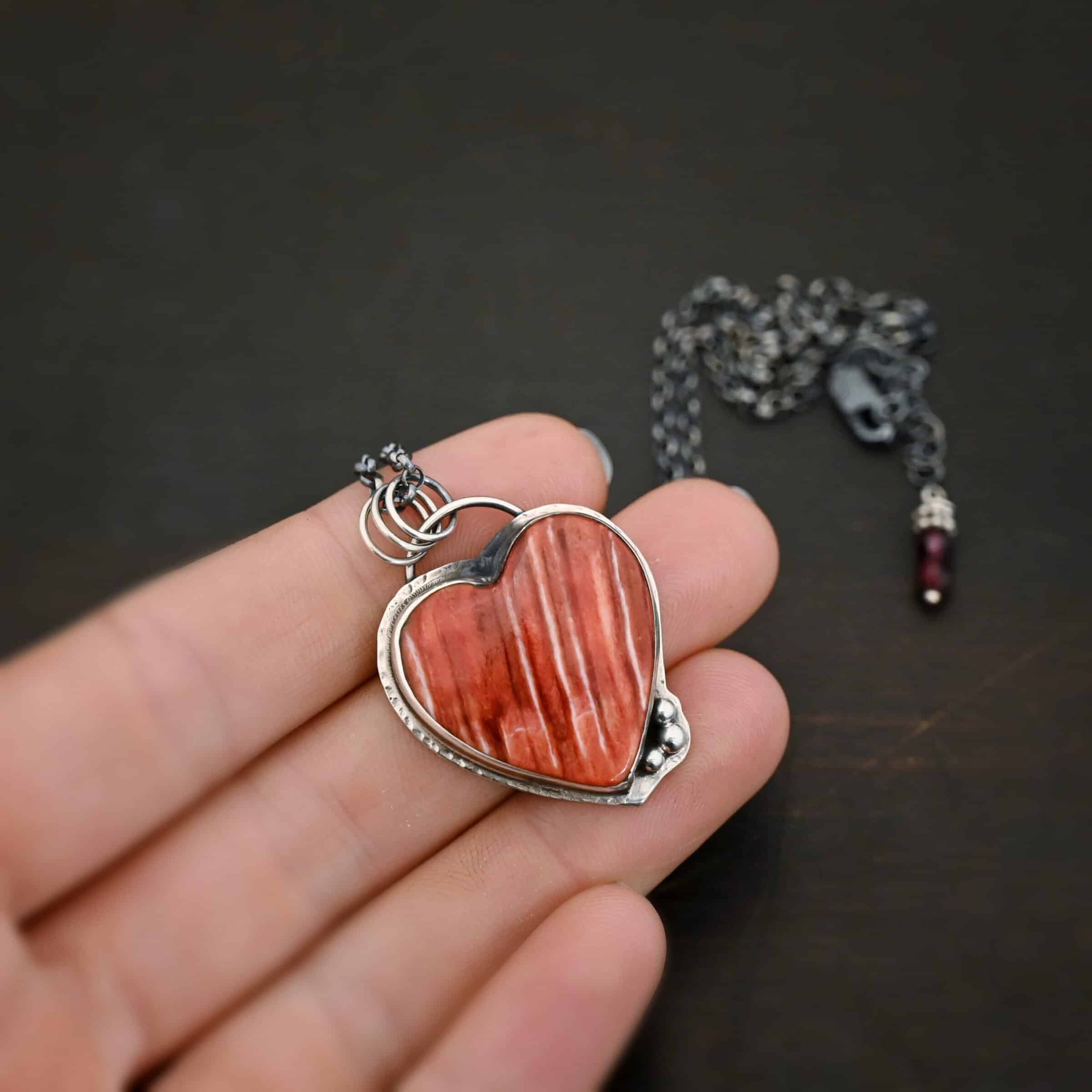Photo of "A Weathered Heart" Necklace. Made out of sterling silver and spiny oyster. The pendant is a heart shaped spiny oyster cabochon. It is a red/orange color mix with some white lines going through it. There are three small silver balls on the bottom right side of the heart pendant. The two outermost balls are smaller, and the ball in the center is larger. They are placed right next to one another, and are close to the tip of the heart. There is a loop on the back of the pendant with three jump rings attached to it. Going through the jump rings are the chain of the necklace. On the end of the extension for the necklace are two garnet beads. Necklace is laying against the fingertips of a hand to demonstrate size.