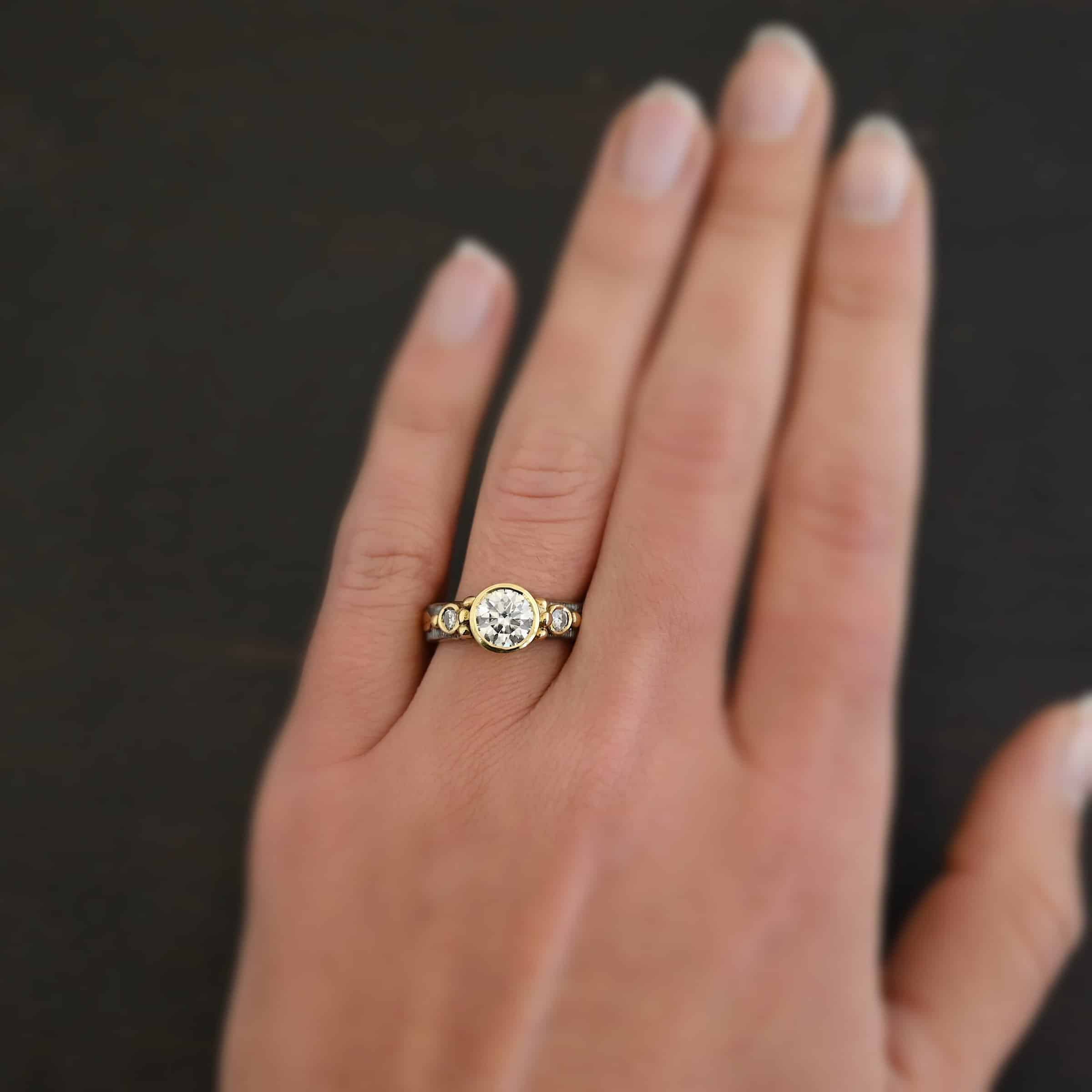 Follow Your Arrow Ring Photo of the "Follow Your Arrow" Ring. Made out of sterling silver, gold, and moissanite. There is a 1.5 carat moissanite in the center of the ring, and two others, one on each side. Each stone is set in gold. Each side of the center moissanite has three gold balls on it, following the curve of the stone setting. Ring is worn on a model's engagement ring finger to demonstrate size.