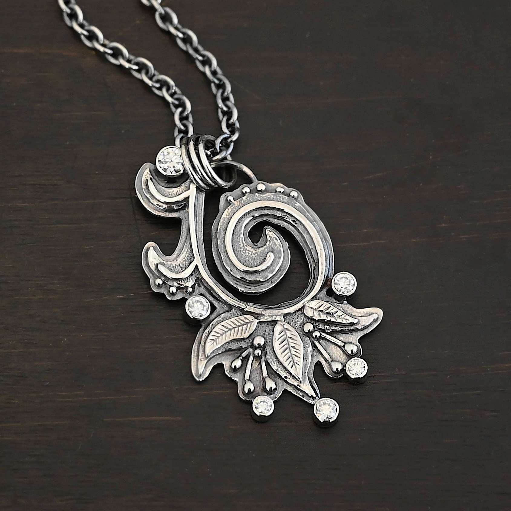 White Lace Necklace 2 Photo of the "White Lace" Necklace. Made out of sterling silver and moissanites. There are six moissanites scattered around the outside edge of the pendant. The inside of the pendant has a swirl that starts on the outside and curves in towards the inside. Attached around the swirl are three silver leaves, along with silver dots and small lines of silver with dots on the end of them. Necklace has a cross and angel wing pendant on the back of the chain There are three jump rings attached to a loop on the pendant in which the chain goes through. Necklace is laying against a dark background.