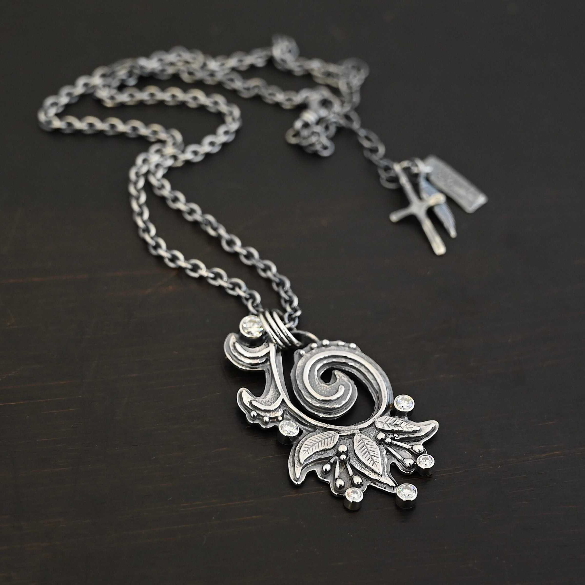 White Lace Necklace 2 Photo of the "White Lace" Necklace. Made out of sterling silver and moissanites. There are six moissanites scattered around the outside edge of the pendant. The inside of the pendant has a swirl that starts on the outside and curves in towards the inside. Attached around the swirl are three silver leaves, along with silver dots and small lines of silver with dots on the end of them. Necklace has a cross and angel wing pendant on the back of the chain There are three jump rings attached to a loop on the pendant in which the chain goes through. Necklace is laying against a dark background.