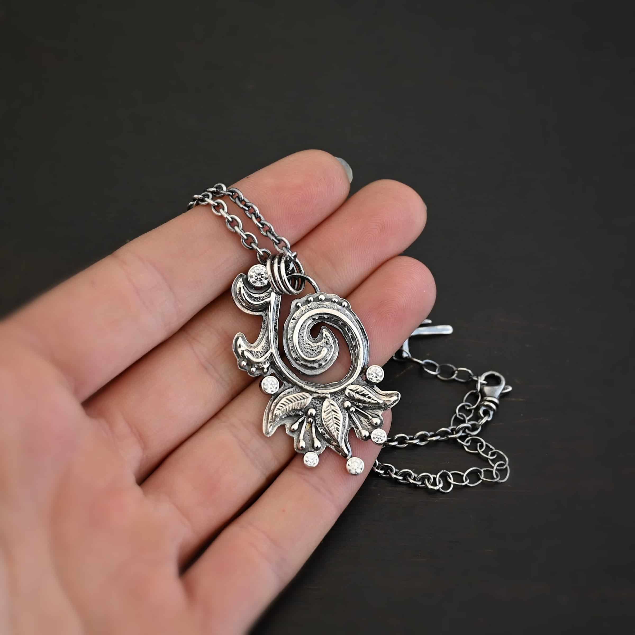 White Lace Necklace 2 Photo of the "White Lace" Necklace. Made out of sterling silver and moissanites. There are six moissanites scattered around the outside edge of the pendant. The inside of the pendant has a swirl that starts on the outside and curves in towards the inside. Attached around the swirl are three silver leaves, along with silver dots and small lines of silver with dots on the end of them. Necklace has a cross and angel wing pendant on the back of the chain There are three jump rings attached to a loop on the pendant in which the chain goes through. Necklace is laying against the fingertips of a hand to demonstrate size.