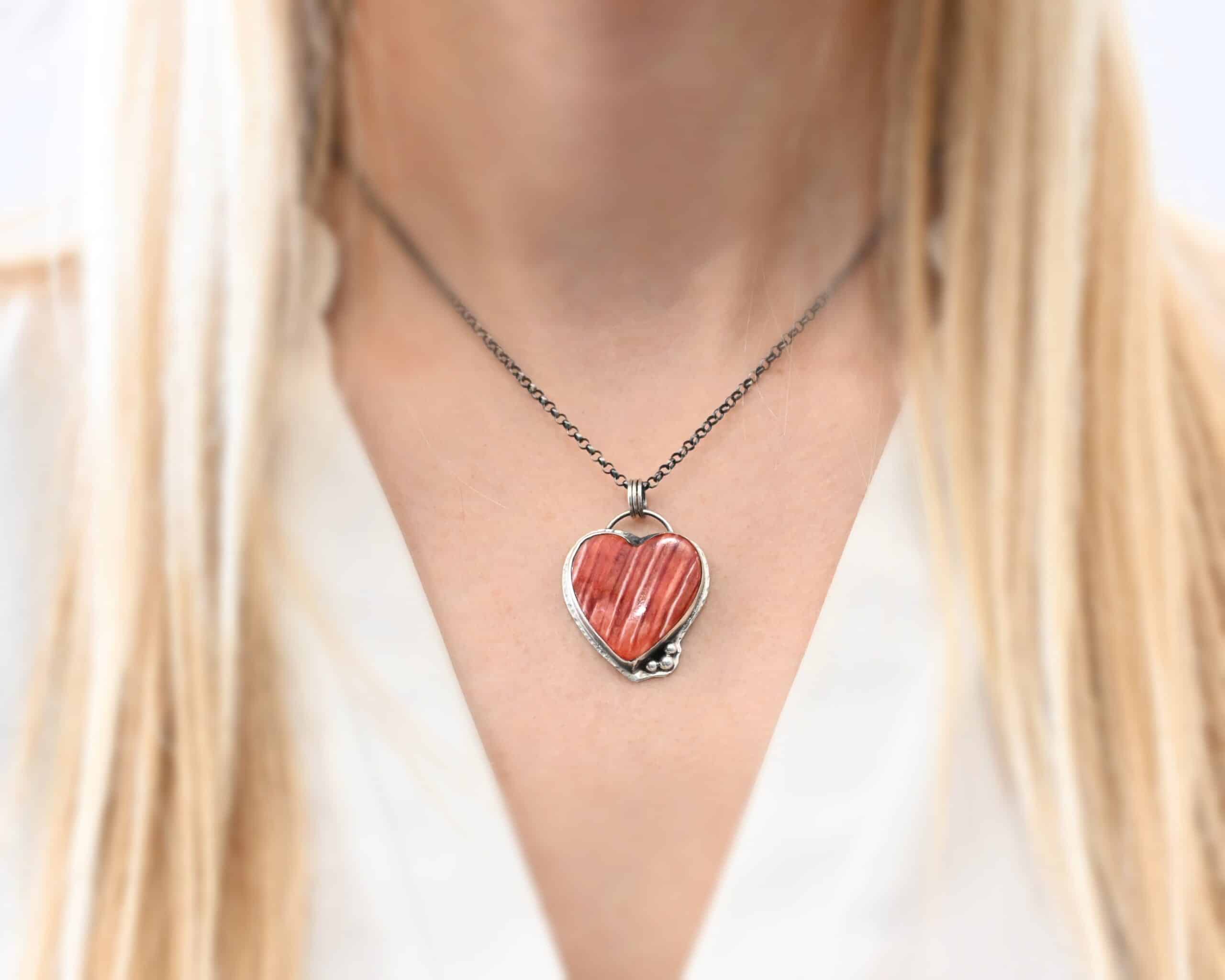 Photo of "A Weathered Heart" Necklace. Made out of sterling silver and spiny oyster. The pendant is a heart shaped spiny oyster cabochon. It is a red/orange color mix with some white lines going through it. There are three small silver balls on the bottom right side of the heart pendant. The two outermost balls are smaller, and the ball in the center is larger. They are placed right next to one another, and are close to the tip of the heart. There is a loop on the back of the pendant with three jump rings attached to it. Going through the jump rings are the chain of the necklace. On the end of the extension for the necklace are two garnet beads. Necklace is worn by a model at 16 inches long to demonstrate size and length. Model is wearing a white v-neck shirt.