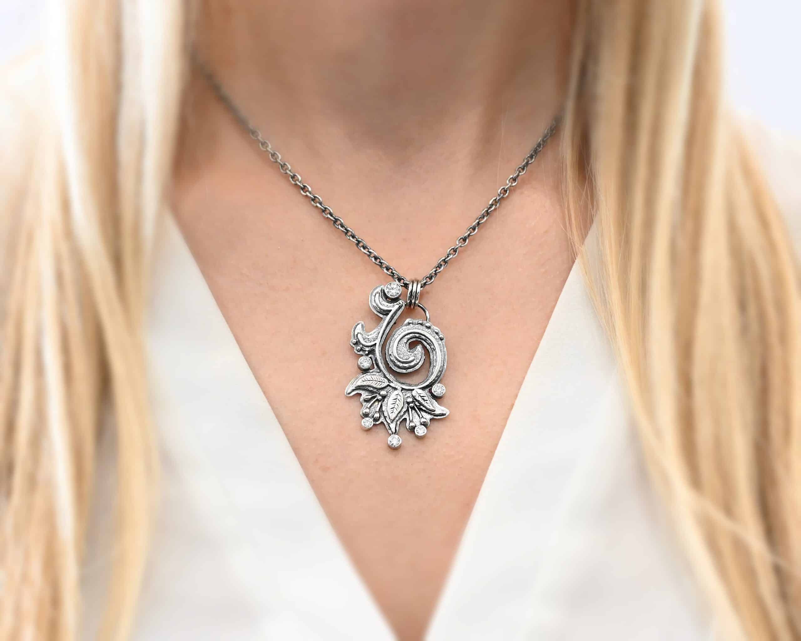 White Lace Necklace Photo of the "White Lace" Necklace. Made out of sterling silver and moissanites. There are six moissanites scattered around the outside edge of the pendant. The inside of the pendant has a swirl that starts on the outside and curves in towards the inside. Attached around the swirl are three silver leaves, along with silver dots and small lines of silver with dots on the end of them. Necklace has a cross and angel wing pendant on the back of the chain There are three jump rings attached to a loop on the pendant in which the chain goes through. Necklace is worn on a model at 16 inches long to demonstrate size and length. Model is wearing a white v-neck shirt.