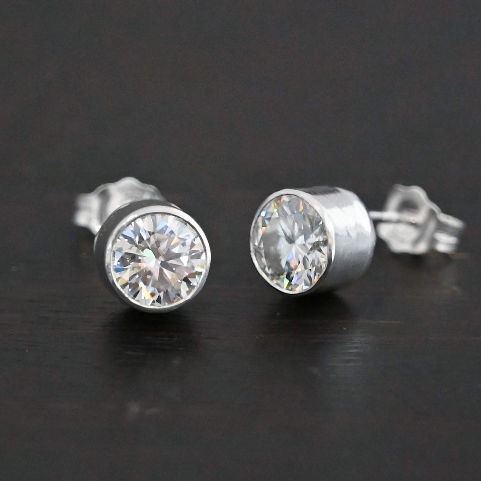 Photo of the "Betina" Earrings. Made out of sterling silver and moissanites. Each earring has a moissanite in the center that is set in sterling silver. Earrings are laying against a dark background.