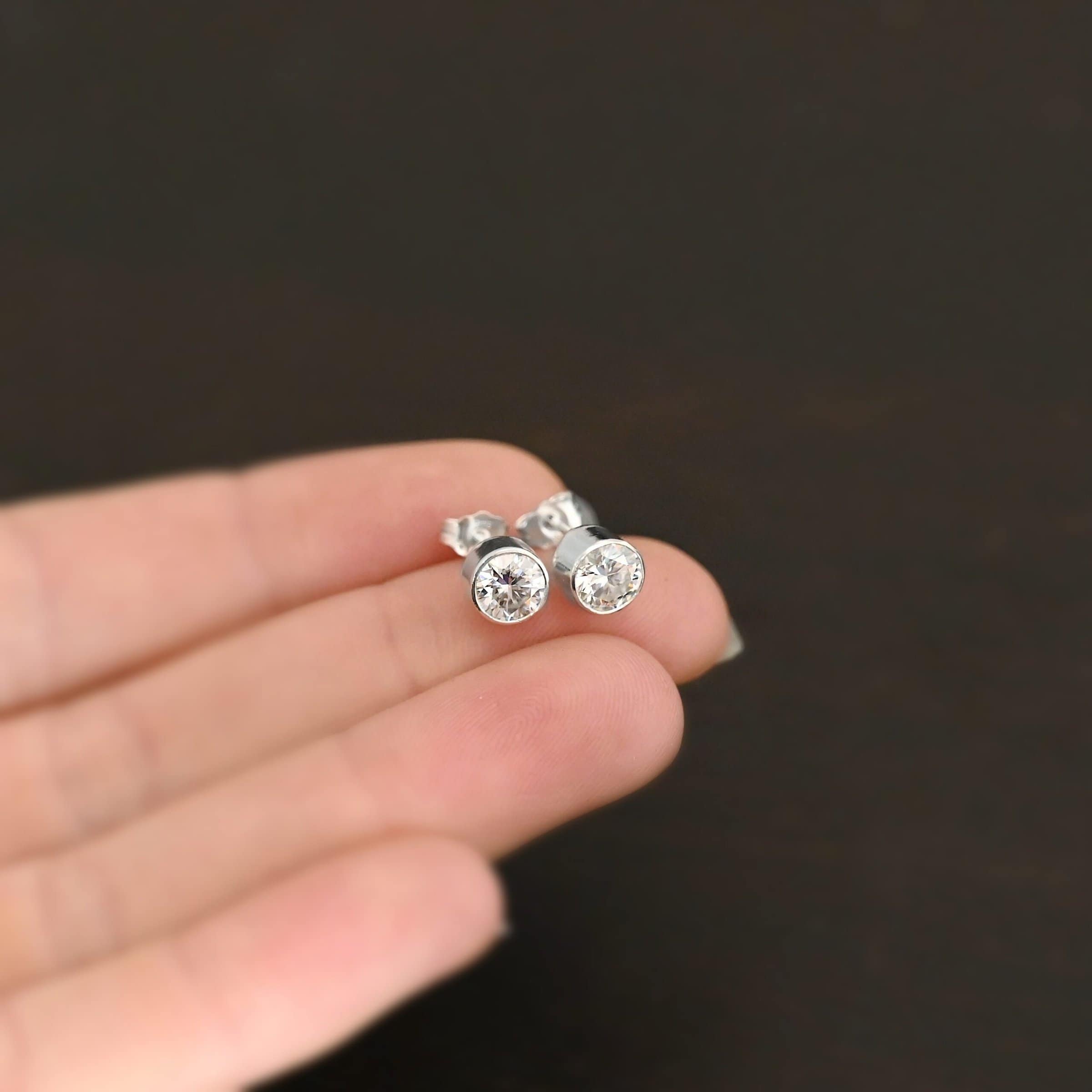 Photo of the "Betina" Earrings. Made out of sterling silver and moissanites. Each earring has a moissanite in the center that is set in sterling silver. Earrings are laying against the fingertips of a models hand to demonstrate size.