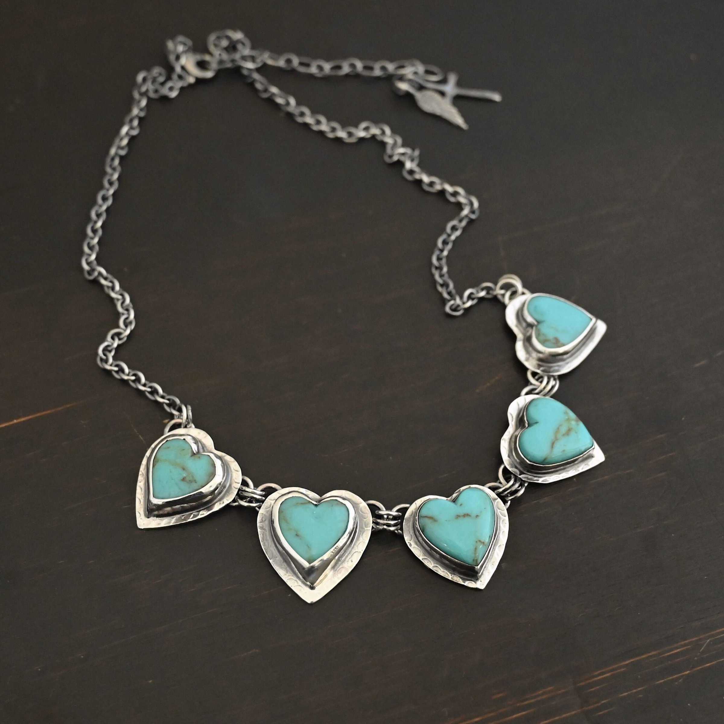 Little Bit of Sweet Necklace Photo of the "Little Bit Of Sweet" Necklace. Made out of sterling silver and turquoise. There are five turquoise hearts, all with two loops on the back. There are three jump rings connected to each loop on the back of the turquoise so all of the hearts are connected to each other. The two outermost hearts have three jump rings that are connected to the chain of the necklace. The chain has a cross and angel wing charm on the back of the necklace. Necklace is laying against a dark background.