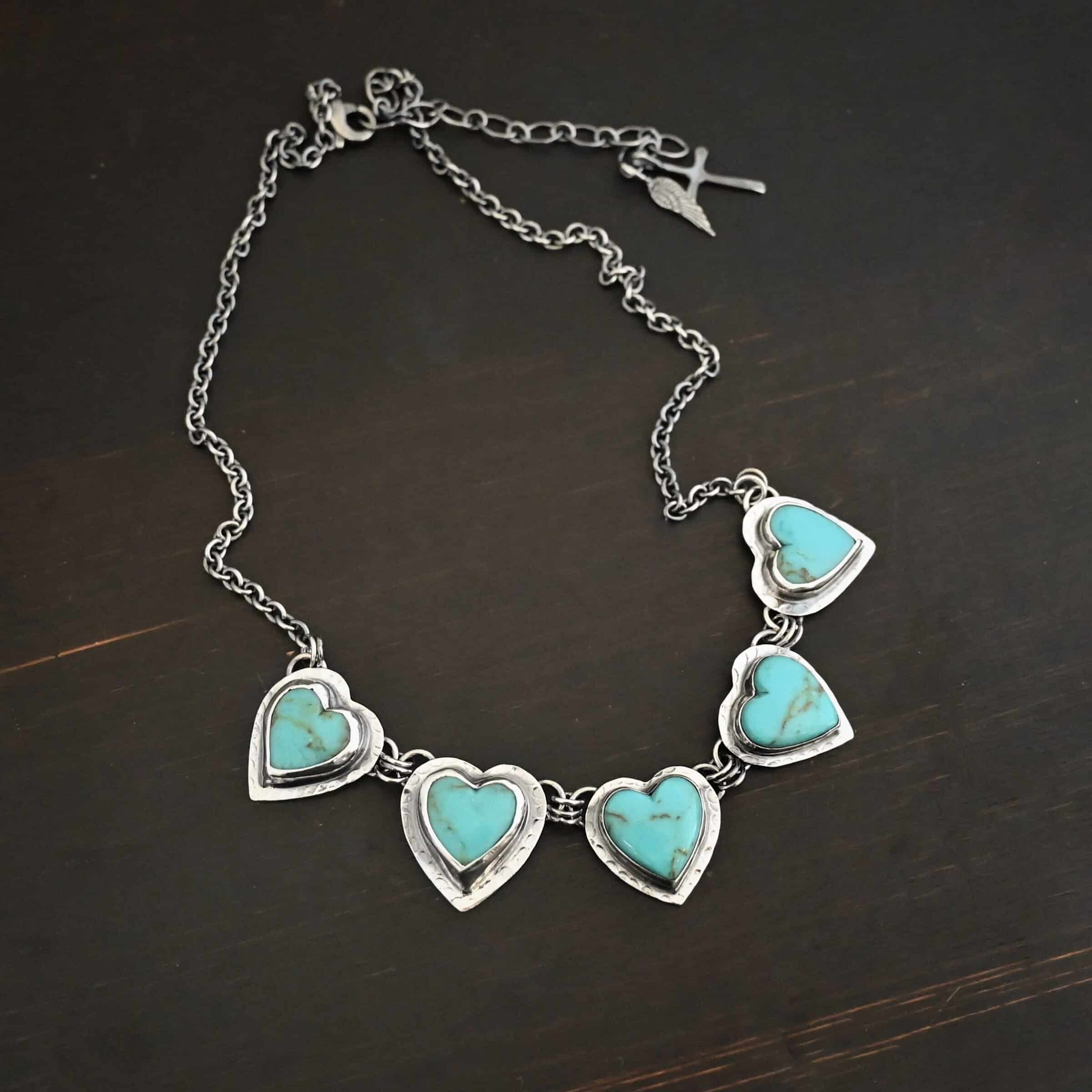 Little Bit of Sweet Necklace Photo of the "Little Bit Of Sweet" Necklace. Made out of sterling silver and turquoise. There are five turquoise hearts, all with two loops on the back. There are three jump rings connected to each loop on the back of the turquoise so all of the hearts are connected to each other. The two outermost hearts have three jump rings that are connected to the chain of the necklace. The chain has a cross and angel wing charm on the back of the necklace. Necklace is laying against a dark background.