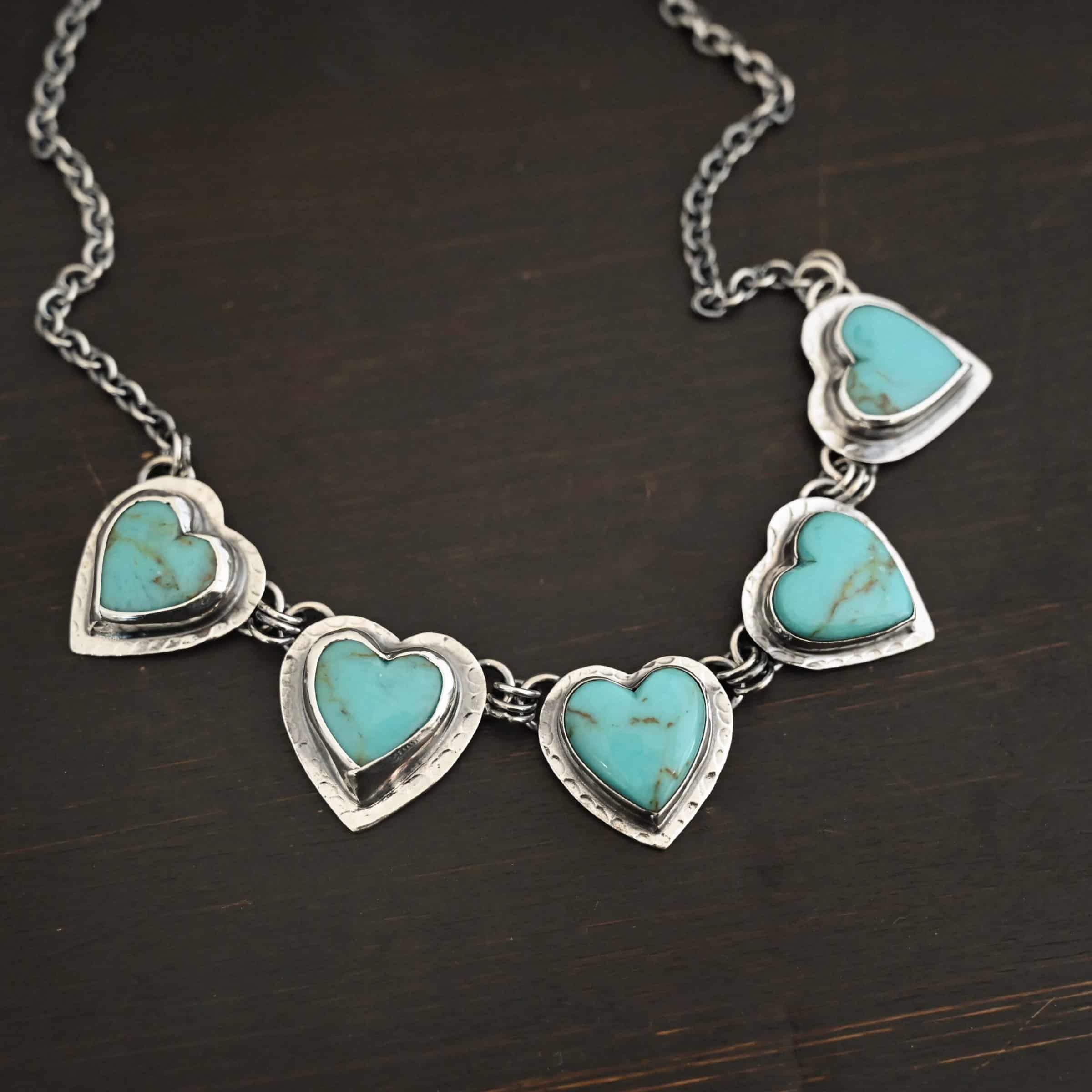Little Bit of Sweet Necklace Photo of the "Little Bit Of Sweet" Necklace. Made out of sterling silver and turquoise. There are five turquoise hearts, all with two loops on the back. There are three jump rings connected to each loop on the back of the turquoise so all of the hearts are connected to each other. The two outermost hearts have three jump rings that are connected to the chain of the necklace. Necklace is laying against a dark background.