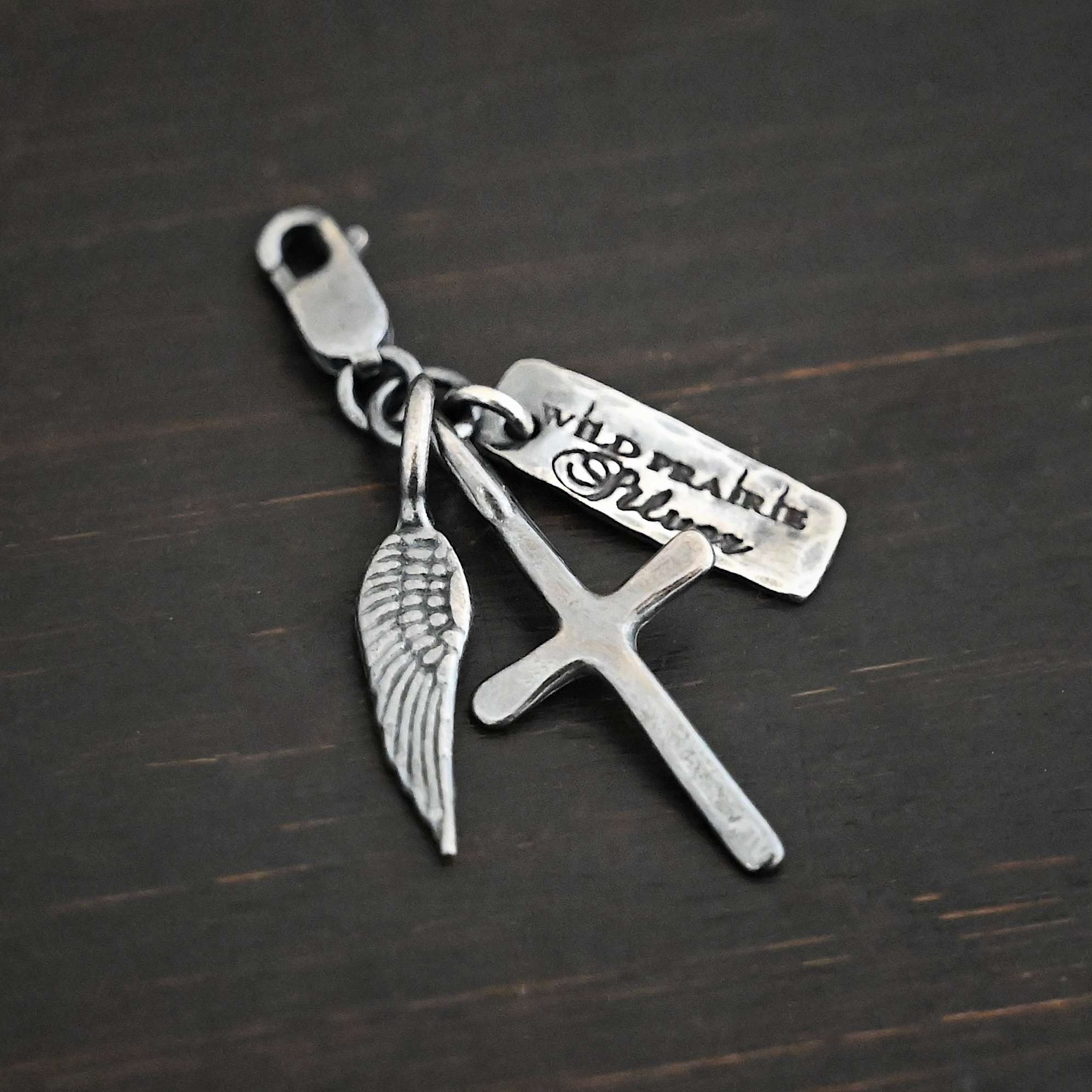 Photo of a cross and angel wing charm. Both are silver. The cross is small and oxidized with a loop on the top of it. The angel wing is oxidized, but also small and has a loop on the top. There is a small rectangle tag that is oxidized and says "Wild Prairie Silver" on it that is colored in dark. All three of these charms are connected to another loop with a lobster claw clasp on the top of the loop. All three are laying against a dark background.