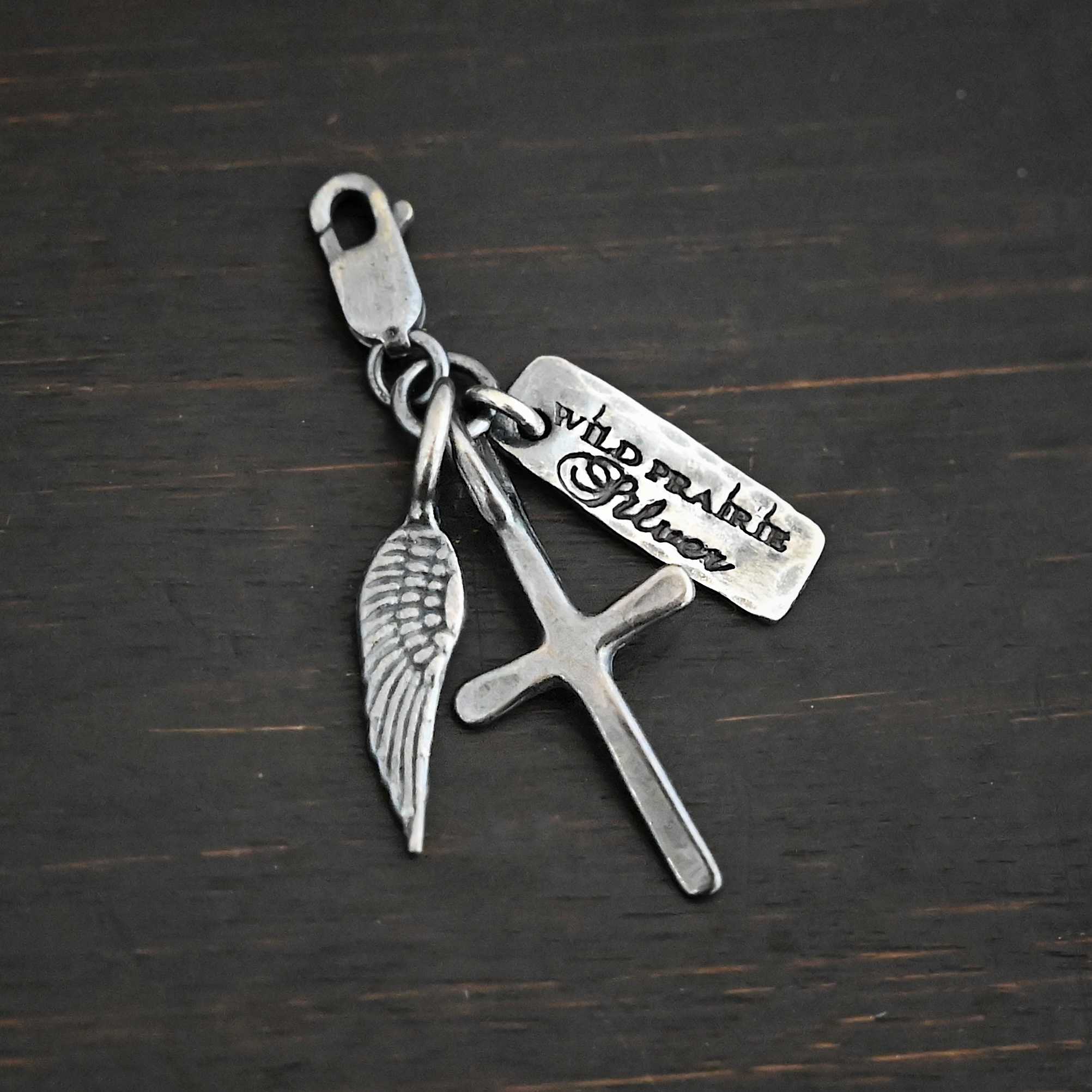 Photo of a cross and angel wing charm. Both are silver. The cross is small and oxidized with a loop on the top of it. The angel wing is oxidized, but also small and has a loop on the top. There is a small rectangle tag that is oxidized and says "Wild Prairie Silver" on it that is colored in dark. All three of these charms are connected to another loop with a lobster claw clasp on the top of the loop. All three are laying against a dark background.