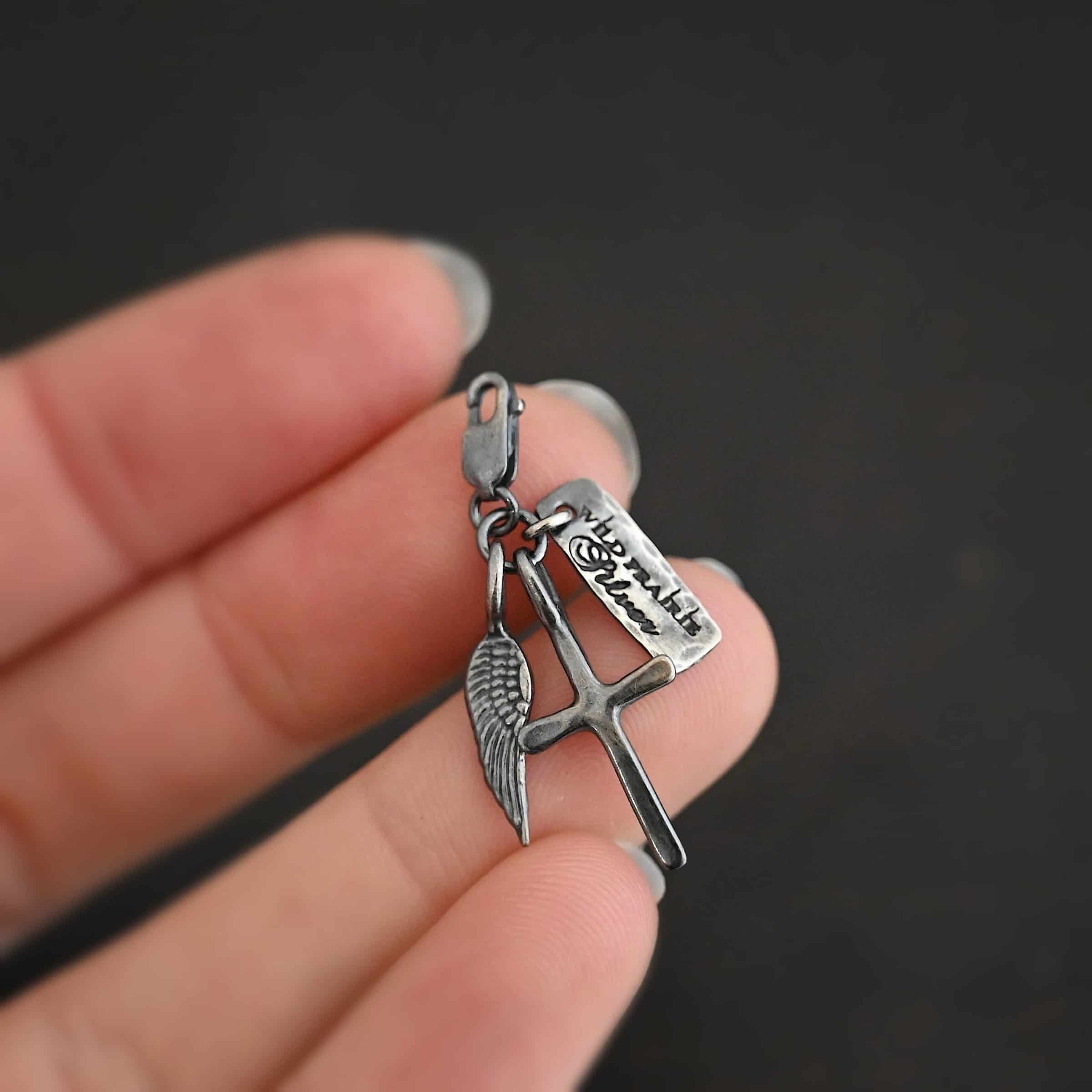 Photo of a cross and angel wing charm. Both are silver. The cross is small and oxidized with a loop on the top of it. The angel wing is oxidized, but also small and has a loop on the top. There is a small rectangle tag that is oxidized and says "Wild Prairie Silver" on it that is colored in dark. All three of these charms are connected to another loop with a lobster claw clasp on the top of the loop. Charms are laying against the fingertips of a hand to demonstrate size.