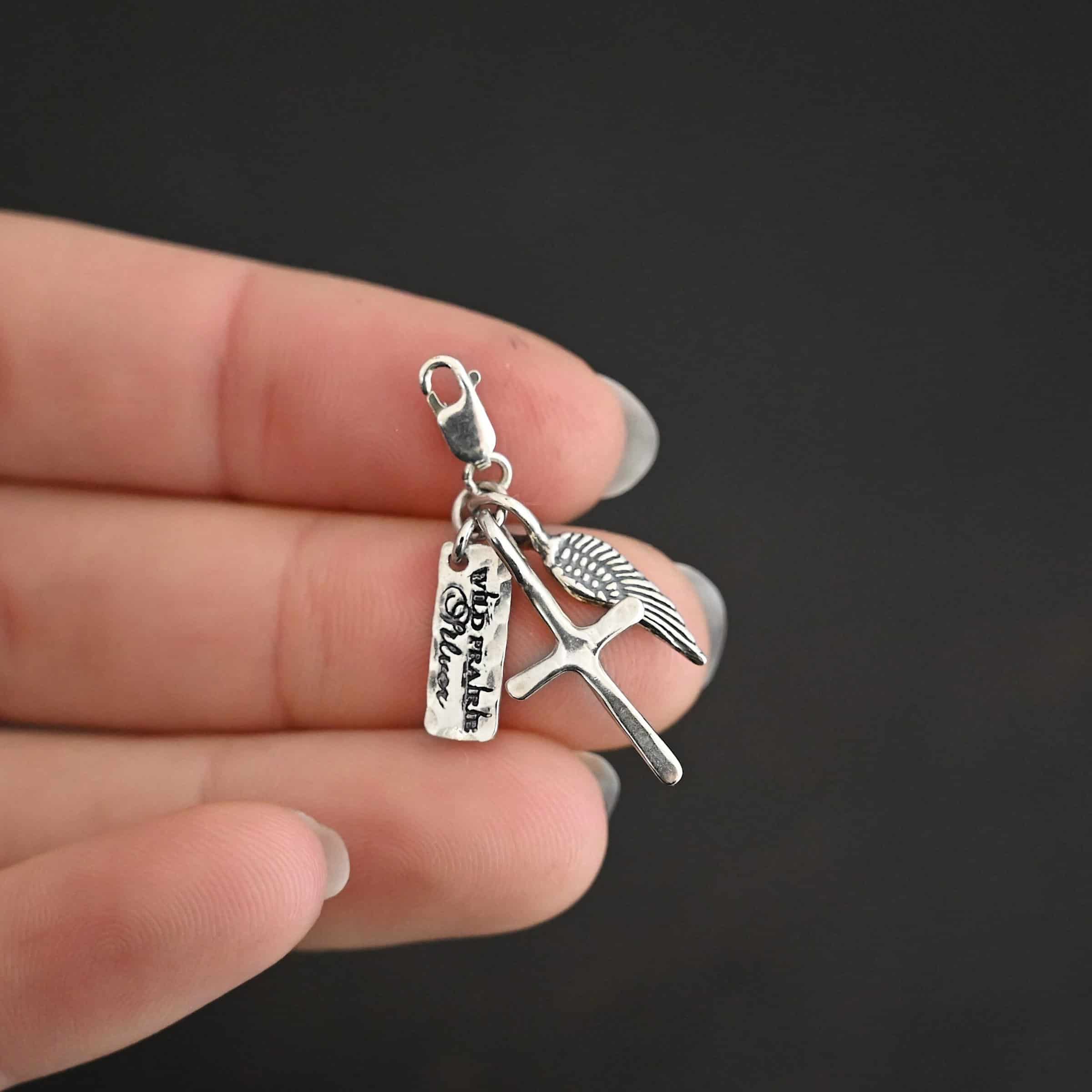Photo of a cross and angel wing charm. Both are silver. The cross is small and shiny with a loop on the top of it. The angel wing is oxidized, but also small and has a loop on the top. There is a small rectangle tag that says "Wild Prairie Silver" on it that is colored in dark. All three of these charms are connected to another loop with a lobster claw clasp on the top of the loop. Charms are laying against the fingertips of a hand to demonstrate size.
