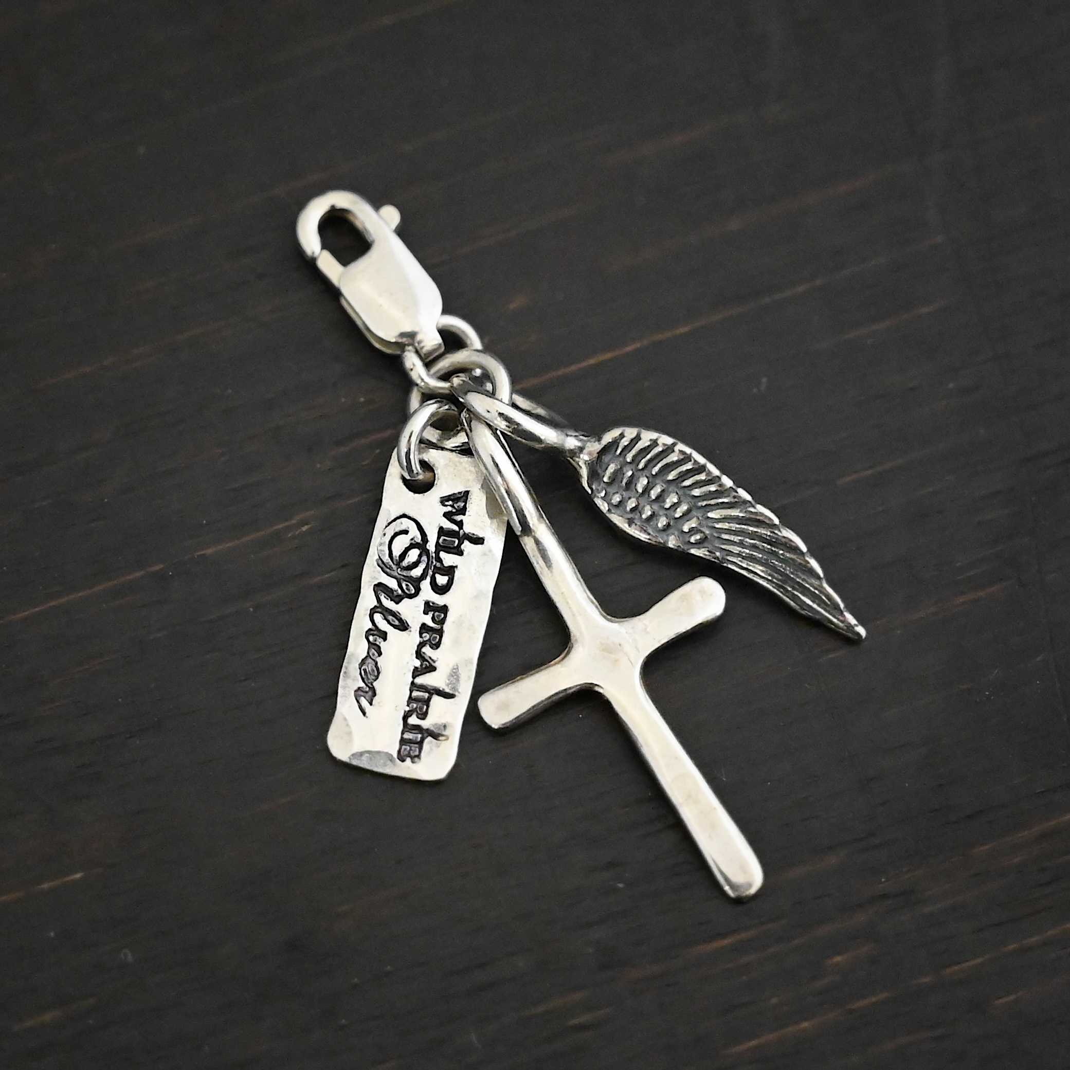 Photo of a cross and angel wing charm. Both are silver. The cross is small and shiny with a loop on the top of it. The angel wing is oxidized, but also small and has a loop on the top. There is a small rectangle tag that says "Wild Prairie Silver" on it that is colored in dark. All three of these charms are connected to another loop with a lobster claw clasp on the top of the loop. All three are laying against a dark background.