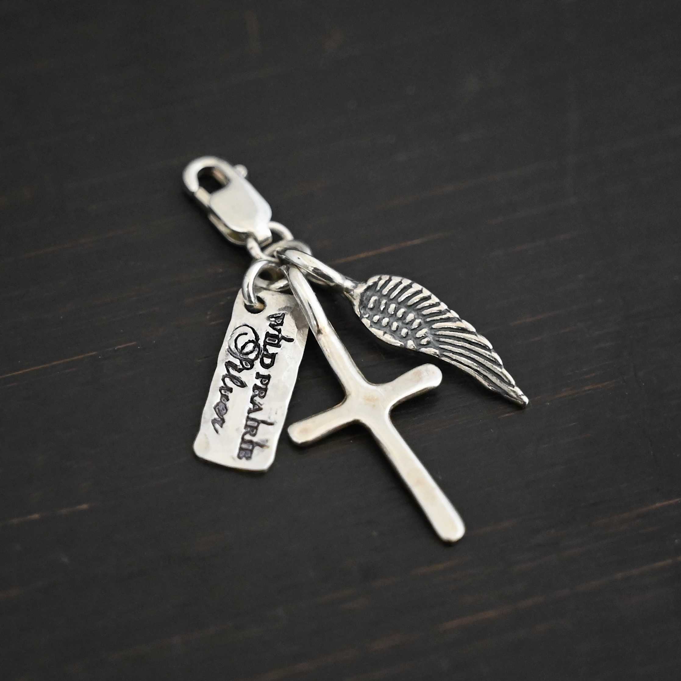 Photo of a cross and angel wing charm. Both are silver. The cross is small and shiny with a loop on the top of it. The angel wing is oxidized, but also small and has a loop on the top. There is a small rectangle tag that says "Wild Prairie Silver" on it that is colored in dark. All three of these charms are connected to another loop with a lobster claw clasp on the top of the loop. All three are laying against a dark background.