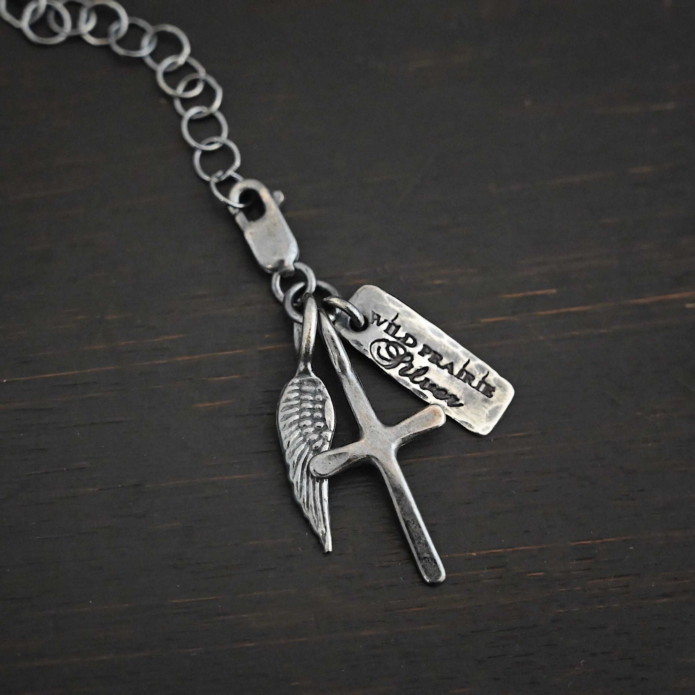 Photo of a cross and angel wing charm. Both are silver. The cross is small and oxidized with a loop on the top of it. The angel wing is oxidized, but also small and has a loop on the top. There is a small rectangle tag that is oxidized and says "Wild Prairie Silver" on it that is colored in dark. All three of these charms are connected to another loop with a lobster claw clasp on the top of the loop. The clasp is attached to a link of sterling silver chain that is oxidized. All three are laying against a dark background.