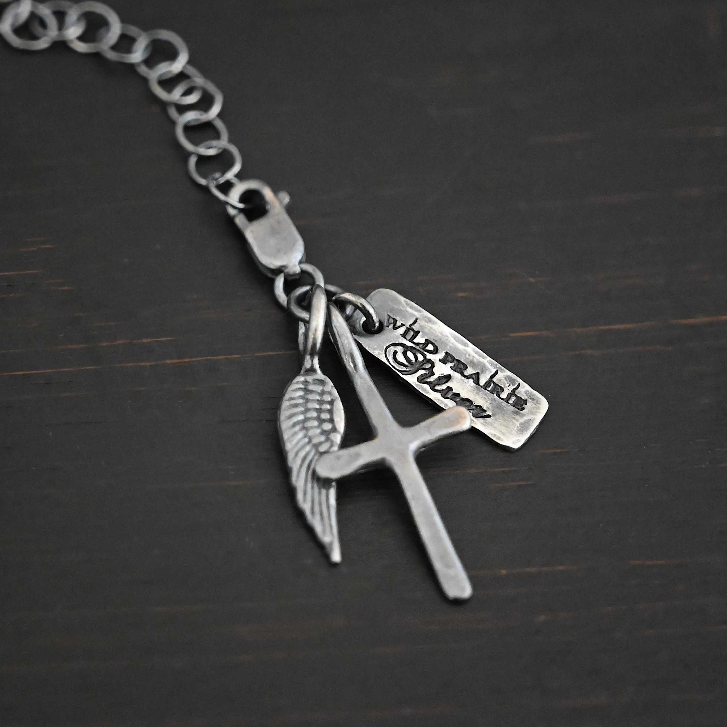 Photo of a cross and angel wing charm. Both are silver. The cross is small and oxidized with a loop on the top of it. The angel wing is oxidized, but also small and has a loop on the top. There is a small rectangle tag that is oxidized and says "Wild Prairie Silver" on it that is colored in dark. All three of these charms are connected to another loop with a lobster claw clasp on the top of the loop. The clasp is attached to a link of sterling silver chain that is oxidized. All three are laying against a dark background.