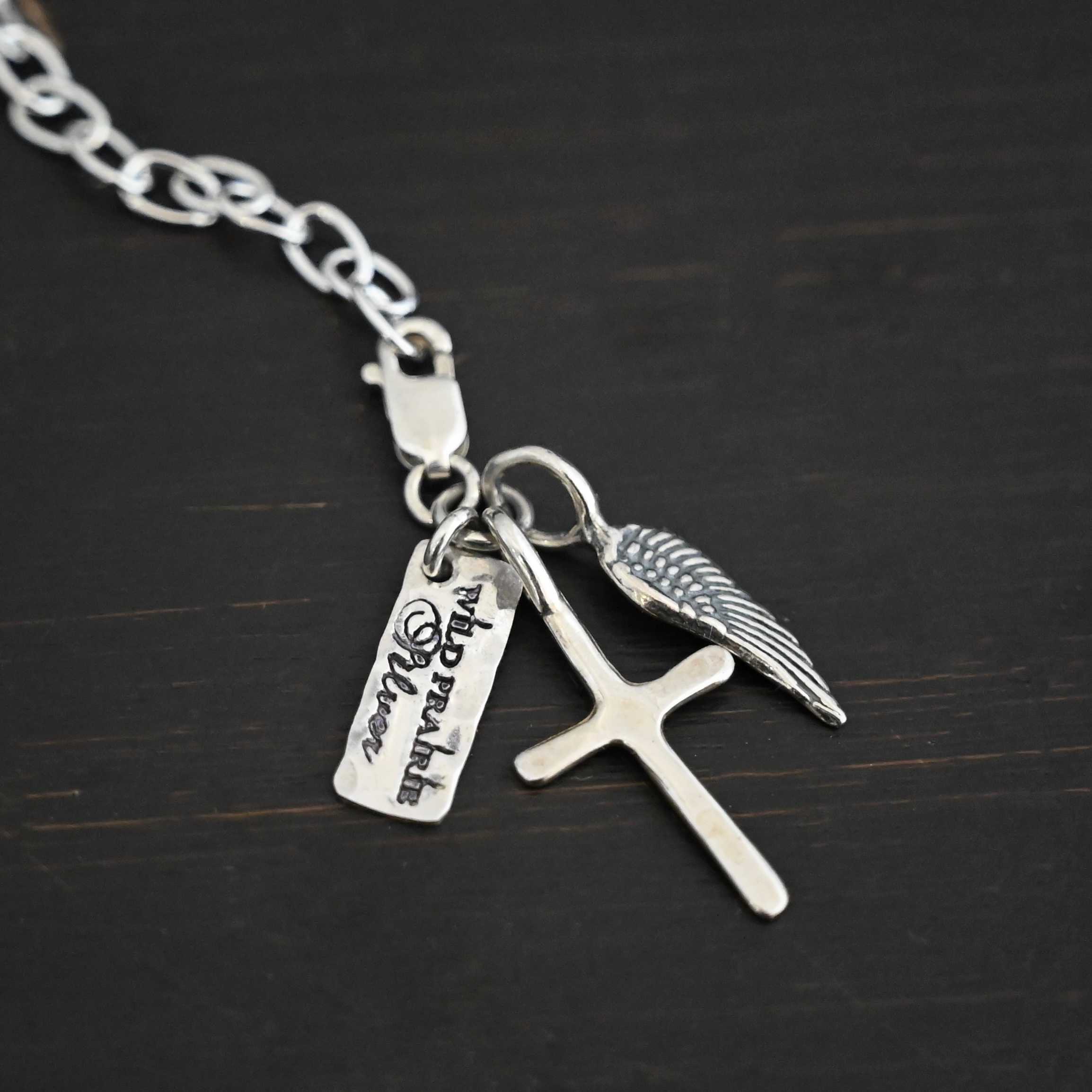 Photo of a cross and angel wing charm. Both are silver. The cross is small and shiny with a loop on the top of it. The angel wing is oxidized, but also small and has a loop on the top. There is a small rectangle tag that says "Wild Prairie Silver" on it that is colored in dark. All three of these charms are connected to another loop with a lobster claw clasp on the top of the loop. The clasp is attached to a link of sterling silver chain. All three are laying against a dark background.
