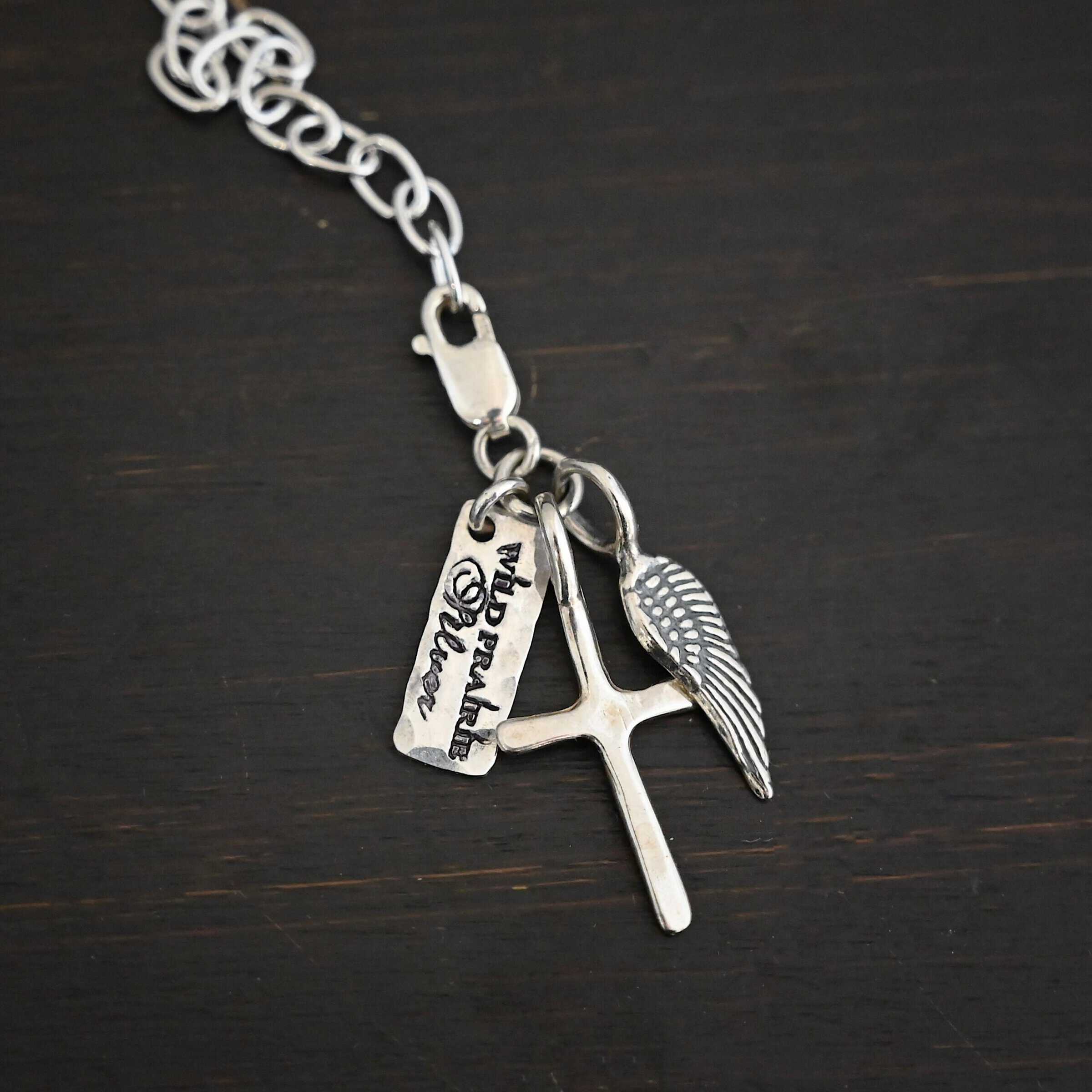 Photo of a cross and angel wing charm. Both are silver. The cross is small and shiny with a loop on the top of it. The angel wing is oxidized, but also small and has a loop on the top. There is a small rectangle tag that says "Wild Prairie Silver" on it that is colored in dark. All three of these charms are connected to another loop with a lobster claw clasp on the top of the loop. The clasp is attached to a link of sterling silver chain. All three are laying against a dark background.