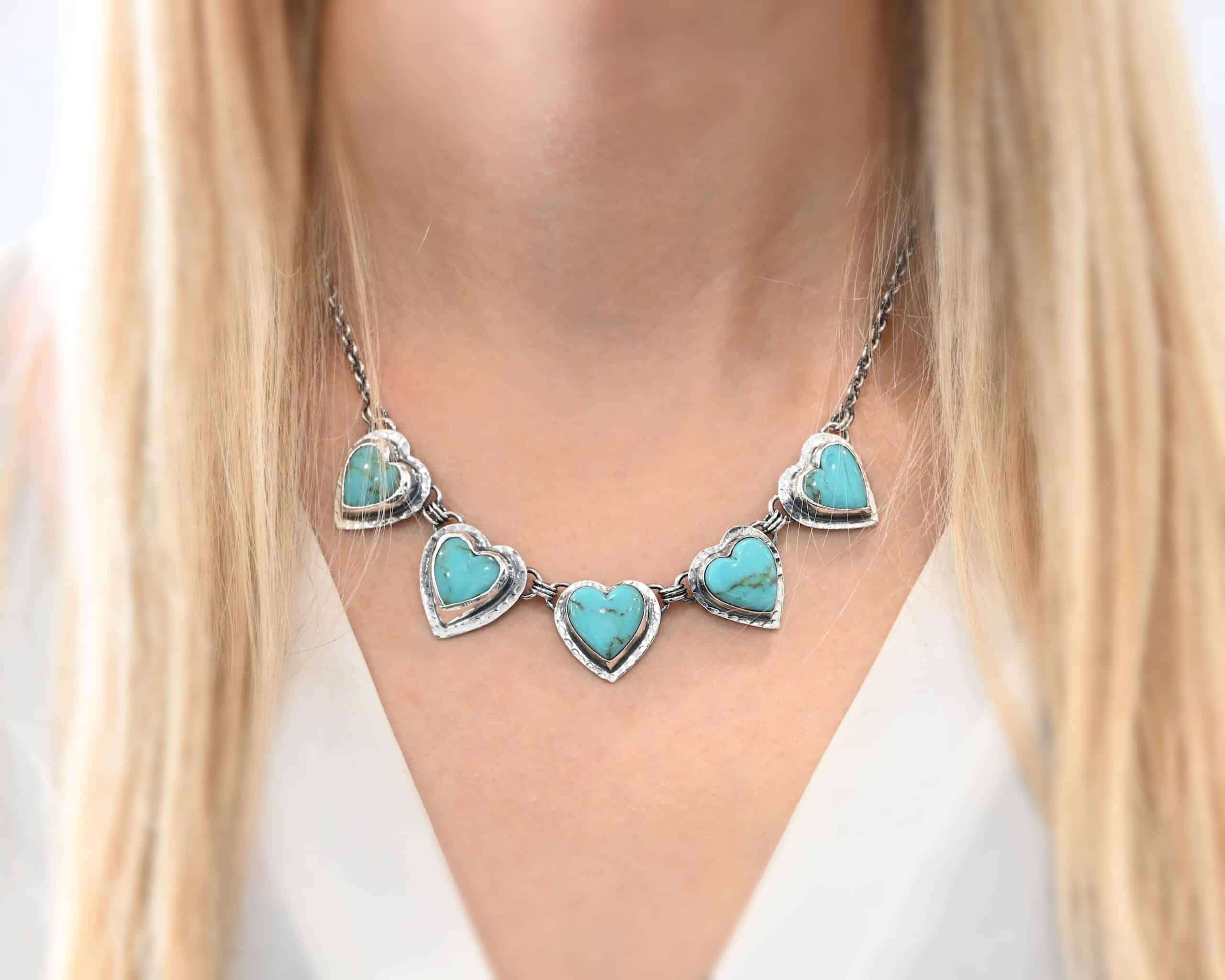 Little Bit of Sweet Necklace Photo of the "Little Bit Of Sweet" Necklace. Made out of sterling silver and turquoise. There are five turquoise hearts, all with two loops on the back. There are three jump rings connected to each loop on the back of the turquoise so all of the hearts are connected to each other. The two outermost hearts have three jump rings that are connected to the chain of the necklace. The chain has a cross and angel wing charm on the back of the necklace. Necklace is worn by a model to demonstrate size.