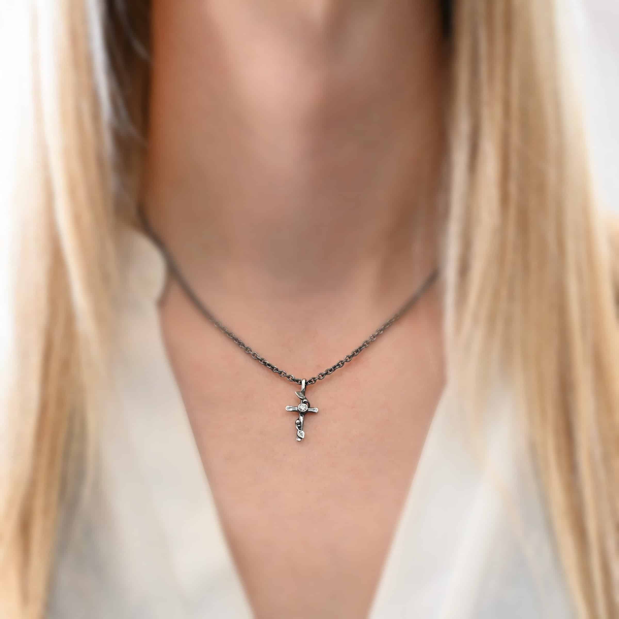 The Third Day Necklace Photo of "The Third Day" Necklace. Made out of sterling silver and a moissanite. The pendant is a sterling silver cross with silver vines and leaves wrapped all around it. There is a small diamond in the center of the cross. There is a loop on top of the cross that is attached to a silver jump ring that are attached to the chain of the necklace. Necklace is worn on a model wearing a white v-neck shirt.