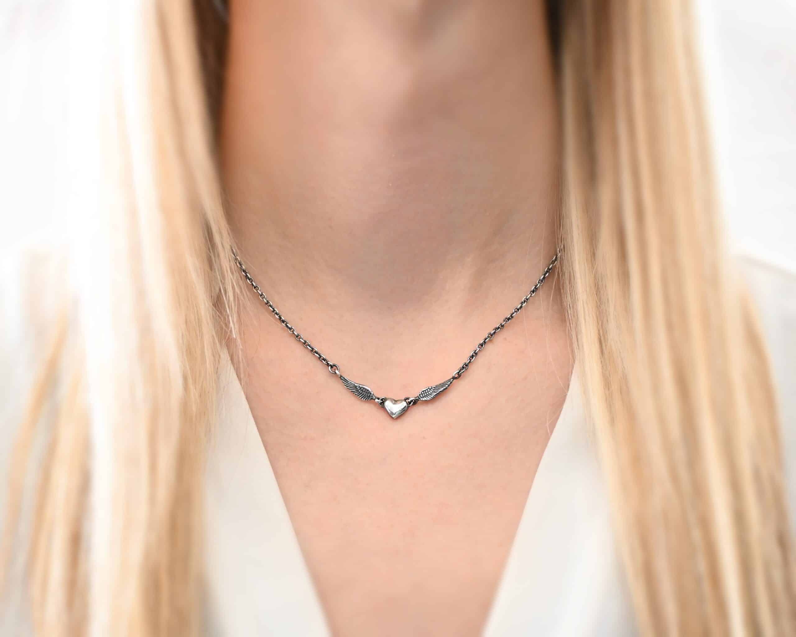 A Little Courage Necklace Photo of "A Little Courage" Necklace. Made out of oxidized sterling silver. There is a small heart pendant in the center of the necklace with a loop on each side of it. Attached to each loop is an angel wing. On the end of each angel wing is a loop that the chain of the necklace is attached to. Necklace is worn by a model wearing a white v-neck shirt.