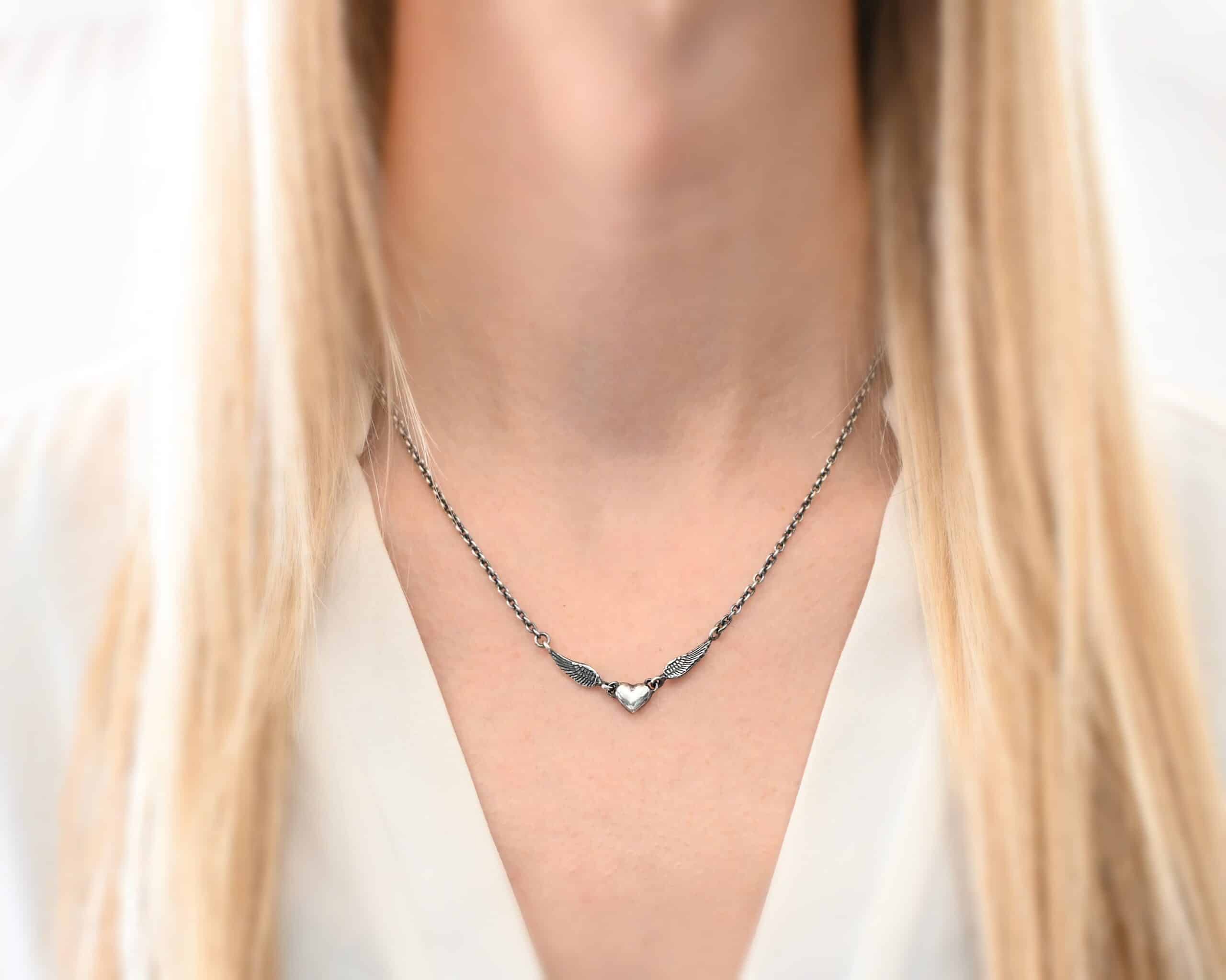 A Little Courage Necklace Photo of "A Little Courage" Necklace. Made out of oxidized sterling silver. There is a small heart pendant in the center of the necklace with a loop on each side of it. Attached to each loop is an angel wing. On the end of each angel wing is a loop that the chain of the necklace is attached to. Necklace is worn by a model wearing a white v-neck shirt.