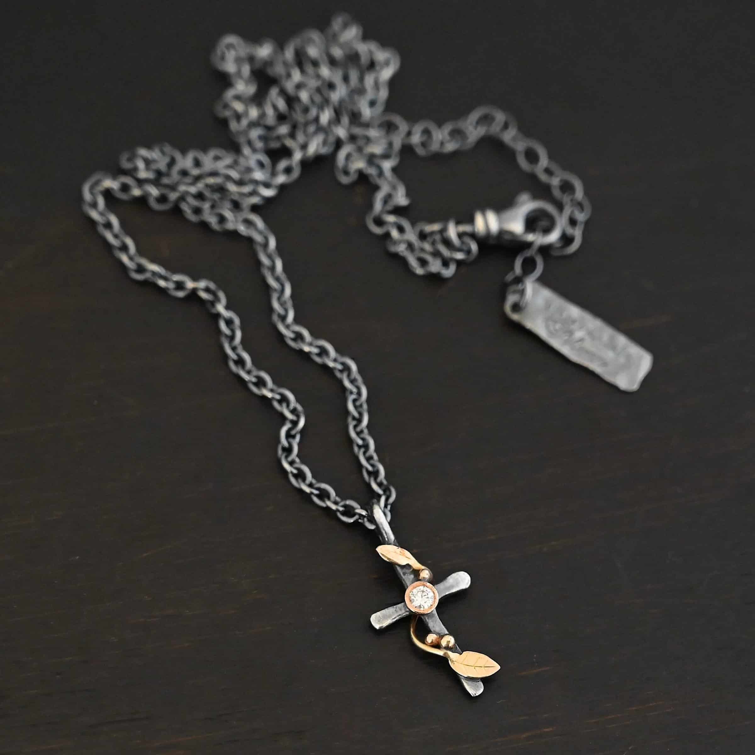 Third Day Necklace Photo of "The Third Day" Necklace. Made out of sterling silver, gold, and a moissanite. The pendant is a sterling silver cross with gold vines and leaves wrapped all around it. There is a small diamond in the center of the cross. There is a loop on top of the cross that is attached to three gold jump rings that are attached to the chain of the necklace. Necklace is laying against a black background.