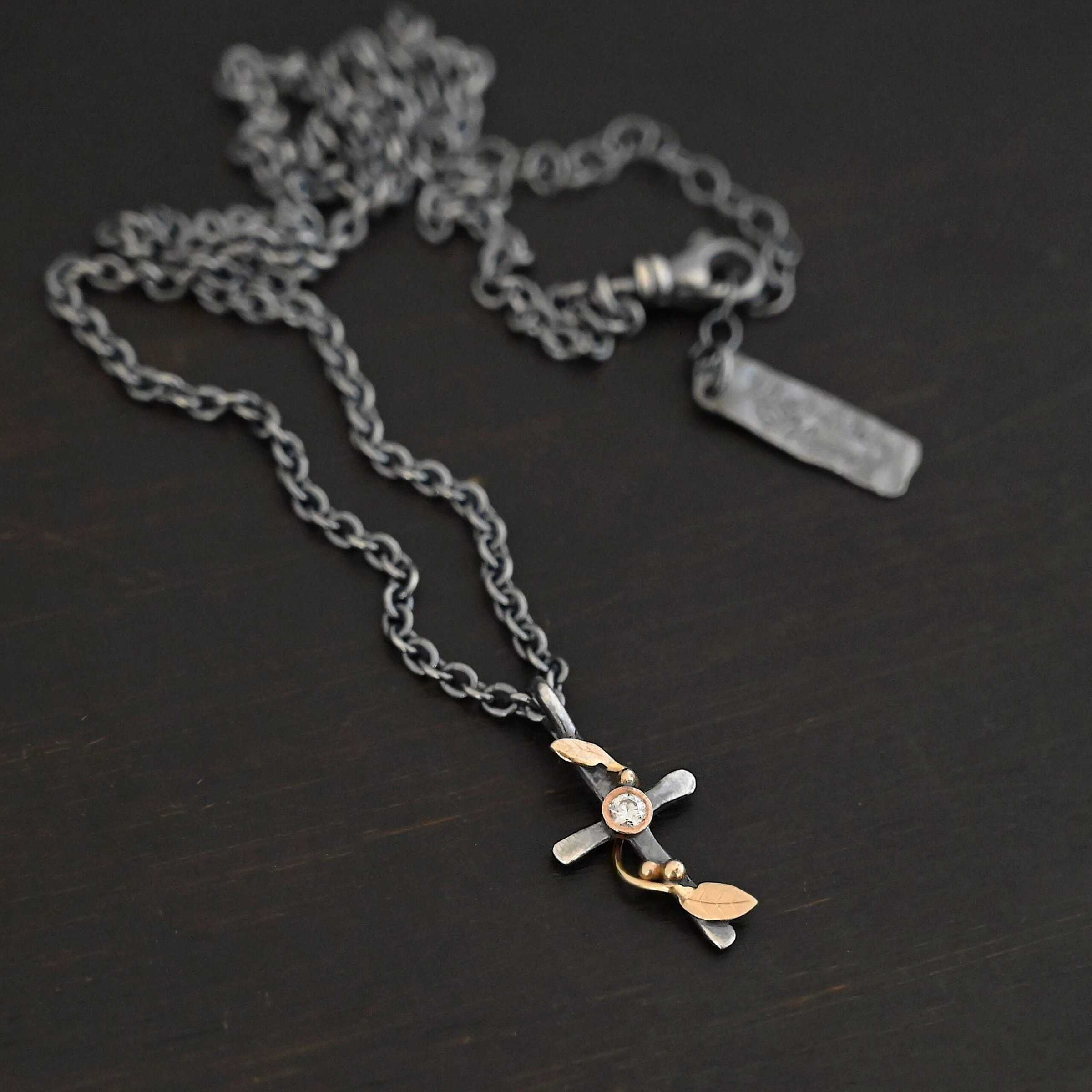 Third Day Necklace Photo of "The Third Day" Necklace. Made out of sterling silver, gold, and a moissanite. The pendant is a sterling silver cross with gold vines and leaves wrapped all around it. There is a small diamond in the center of the cross. There is a loop on top of the cross that is attached to three gold jump rings that are attached to the chain of the necklace. Necklace is laying against a black background.