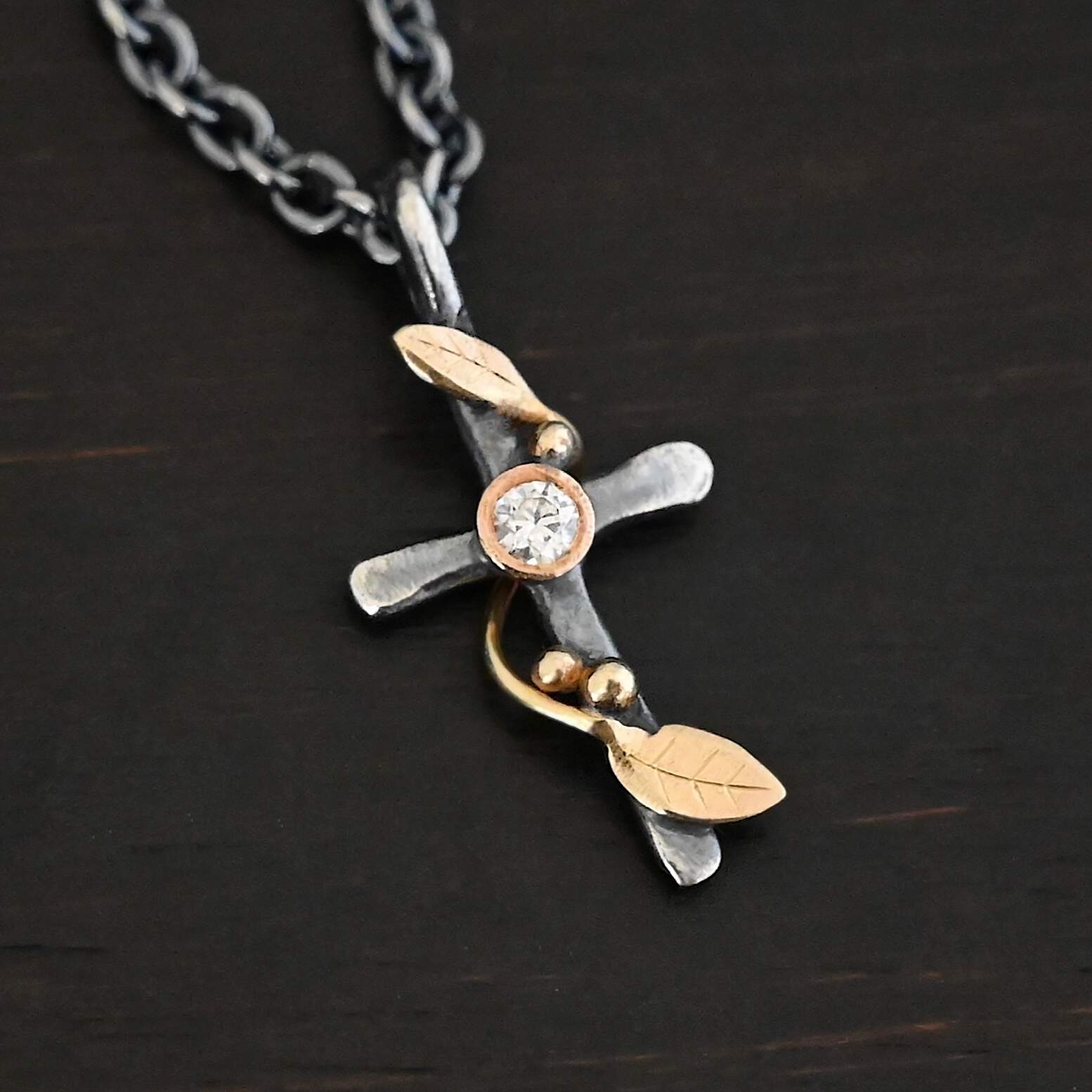 Third Day Necklace Photo of "The Third Day" Necklace. Made out of sterling silver, gold, and a moissanite. The pendant is a sterling silver cross with gold vines and leaves wrapped all around it. There is a small diamond in the center of the cross. There is a loop on top of the cross that is attached to three gold jump rings that are attached to the chain of the necklace. Necklace is laying against a black background.