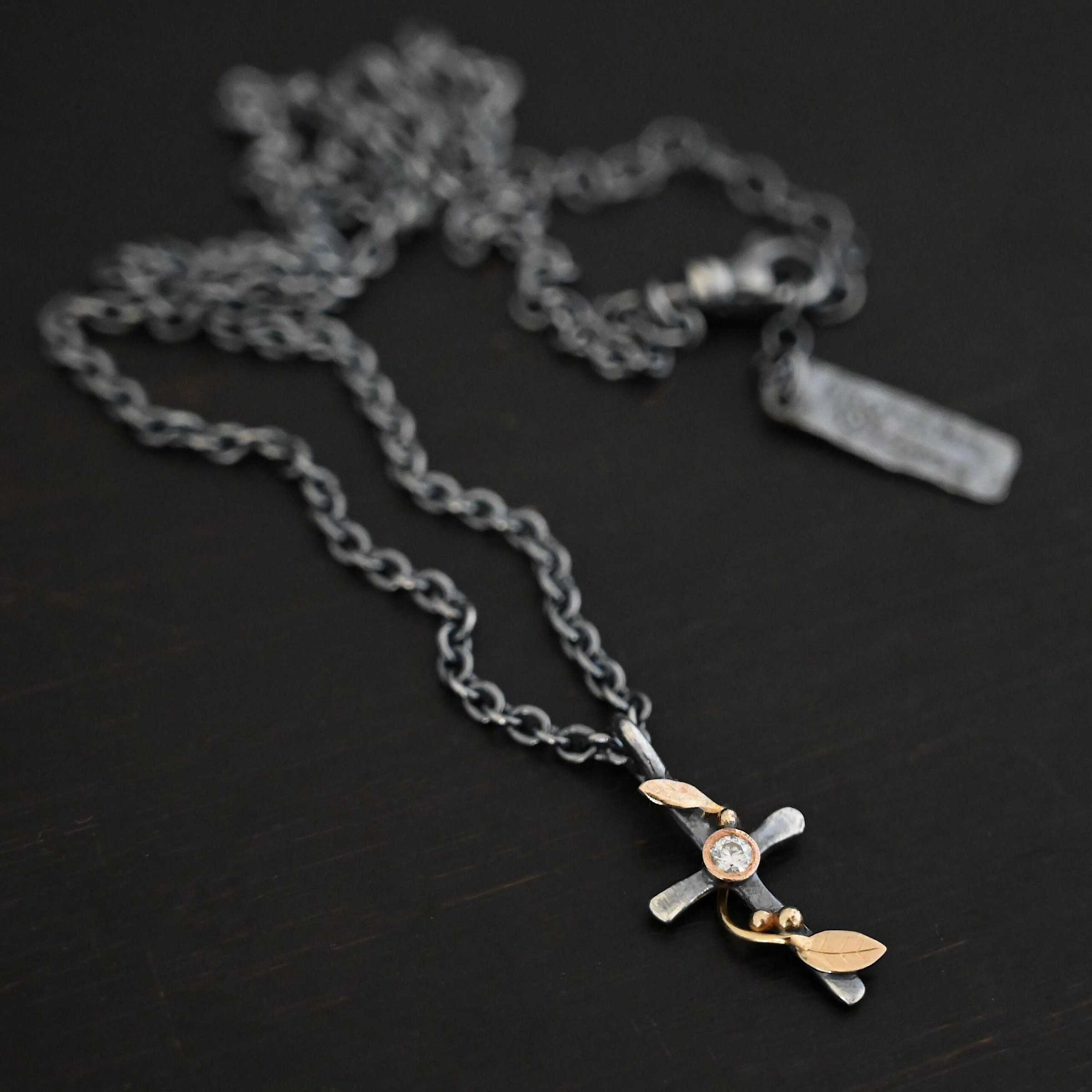 Third Day Necklace Photo of "The Third Day" Necklace. Made out of sterling silver, gold, and a moissanite. The pendant is a sterling silver cross with gold vines and leaves wrapped all around it. There is a small diamond in the center of the cross. There is a loop on top of the cross that is attached to three gold jump rings that are attached to the chain of the necklace. Necklace is laying against a black background.
