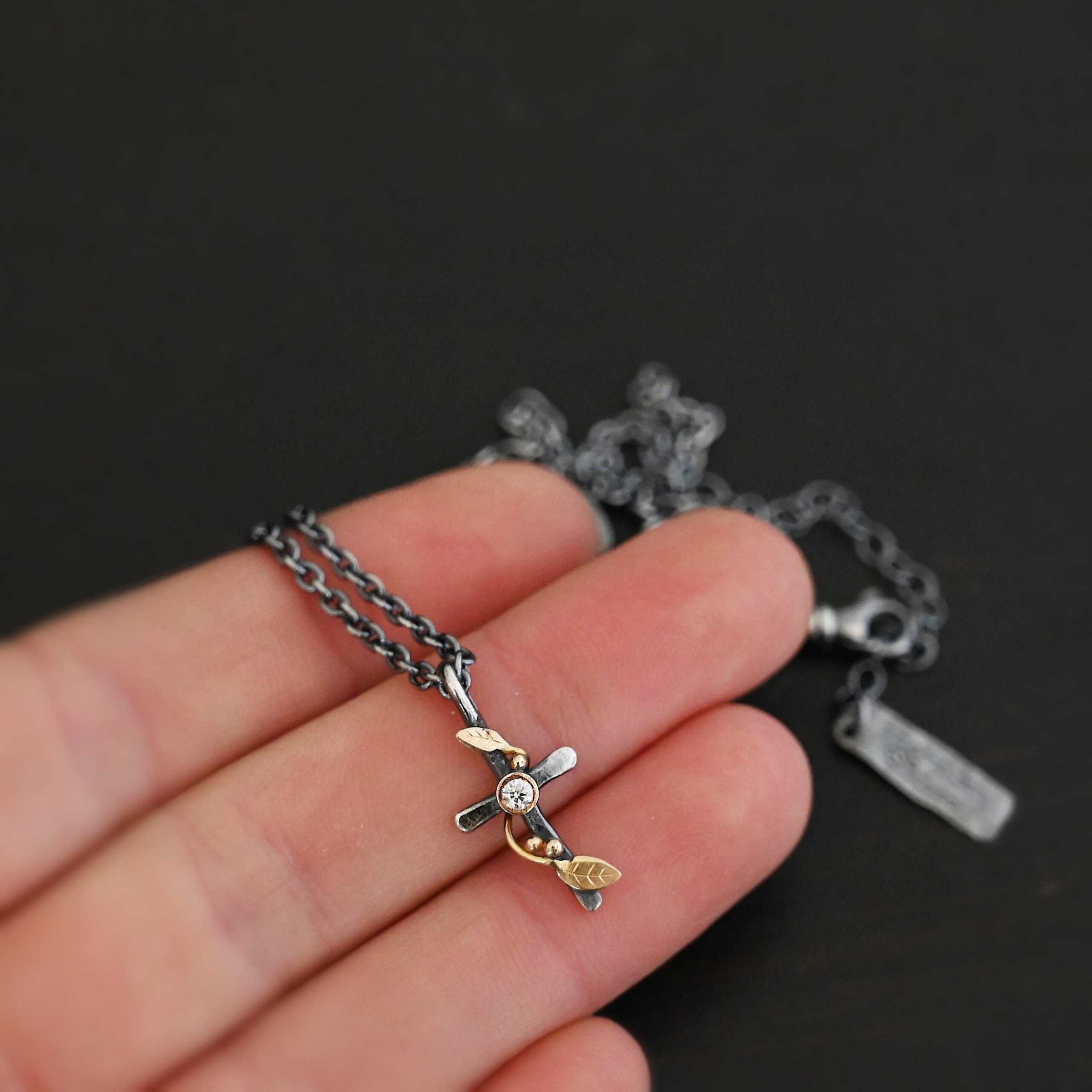Third Day Necklace Photo of "The Third Day" Necklace. Made out of sterling silver, gold, and a diamond. The pendant is a sterling silver cross with gold vines and leaves wrapped all around it. There is a small diamond in the center of the cross. There is a loop on top of the cross that is attached to three gold jump rings that are attached to the chain of the necklace. Necklace is held on the fingertips of a hand to demonstrate size.