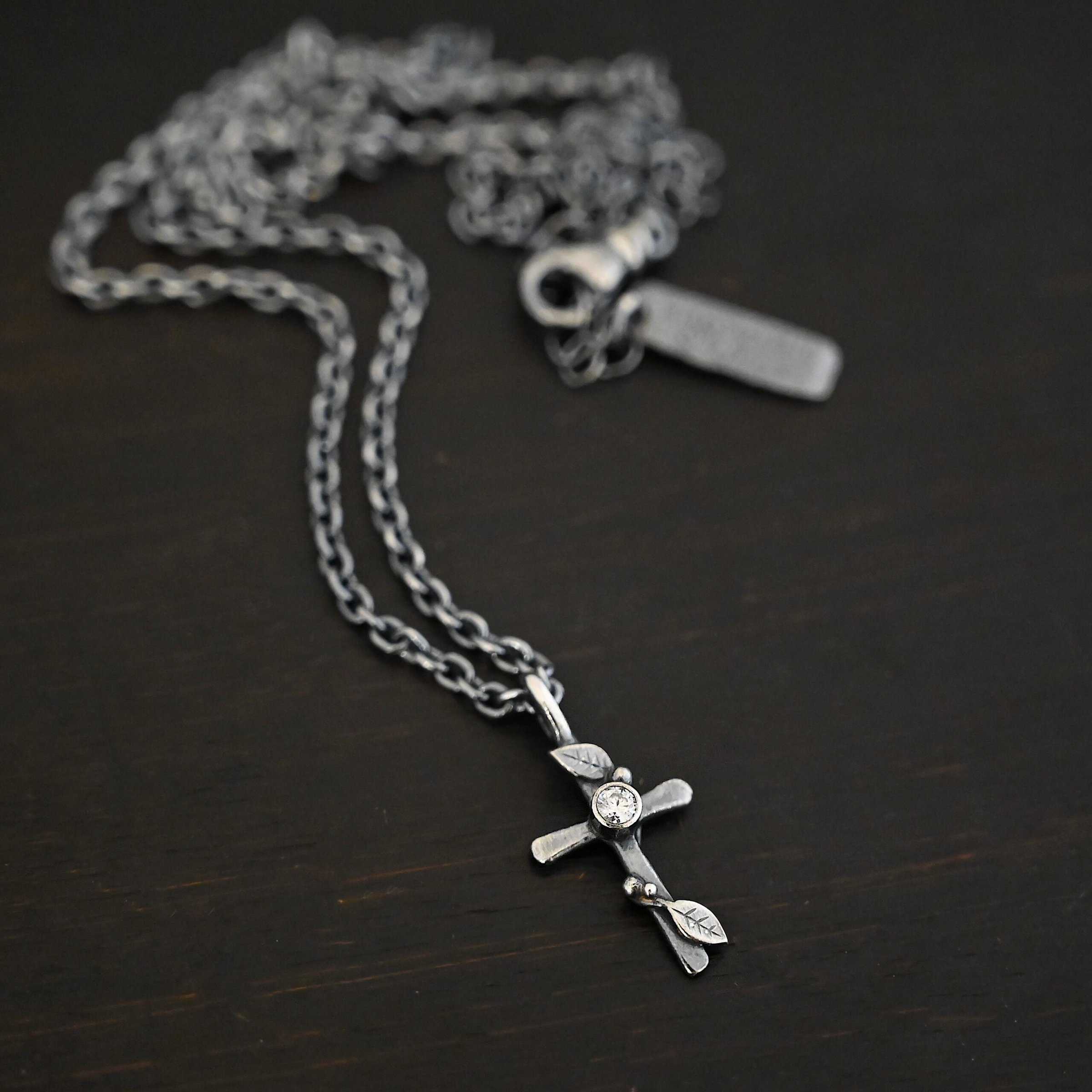 Third Day Necklace Photo of "The Third Day" Necklace. Made out of sterling silver and a moissanite. The pendant is a sterling silver cross with silver vines and leaves wrapped all around it. There is a small diamond in the center of the cross. There is a loop on top of the cross that is attached to a silver jump ring that are attached to the chain of the necklace. Necklace is laying against a black background.