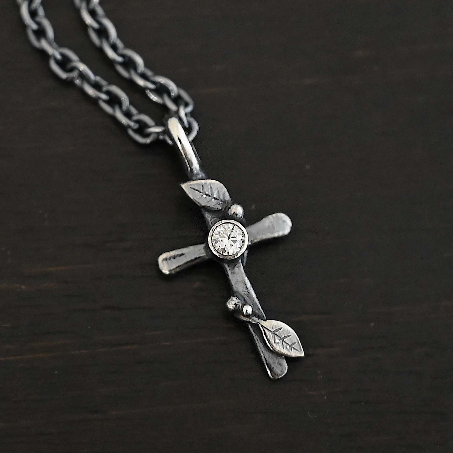 Third Day Necklace Photo of "The Third Day" Necklace. Made out of sterling silver and a moissanite. The pendant is a sterling silver cross with silver vines and leaves wrapped all around it. There is a small diamond in the center of the cross. There is a loop on top of the cross that is attached to a silver jump ring that are attached to the chain of the necklace. Necklace is laying against a black background.