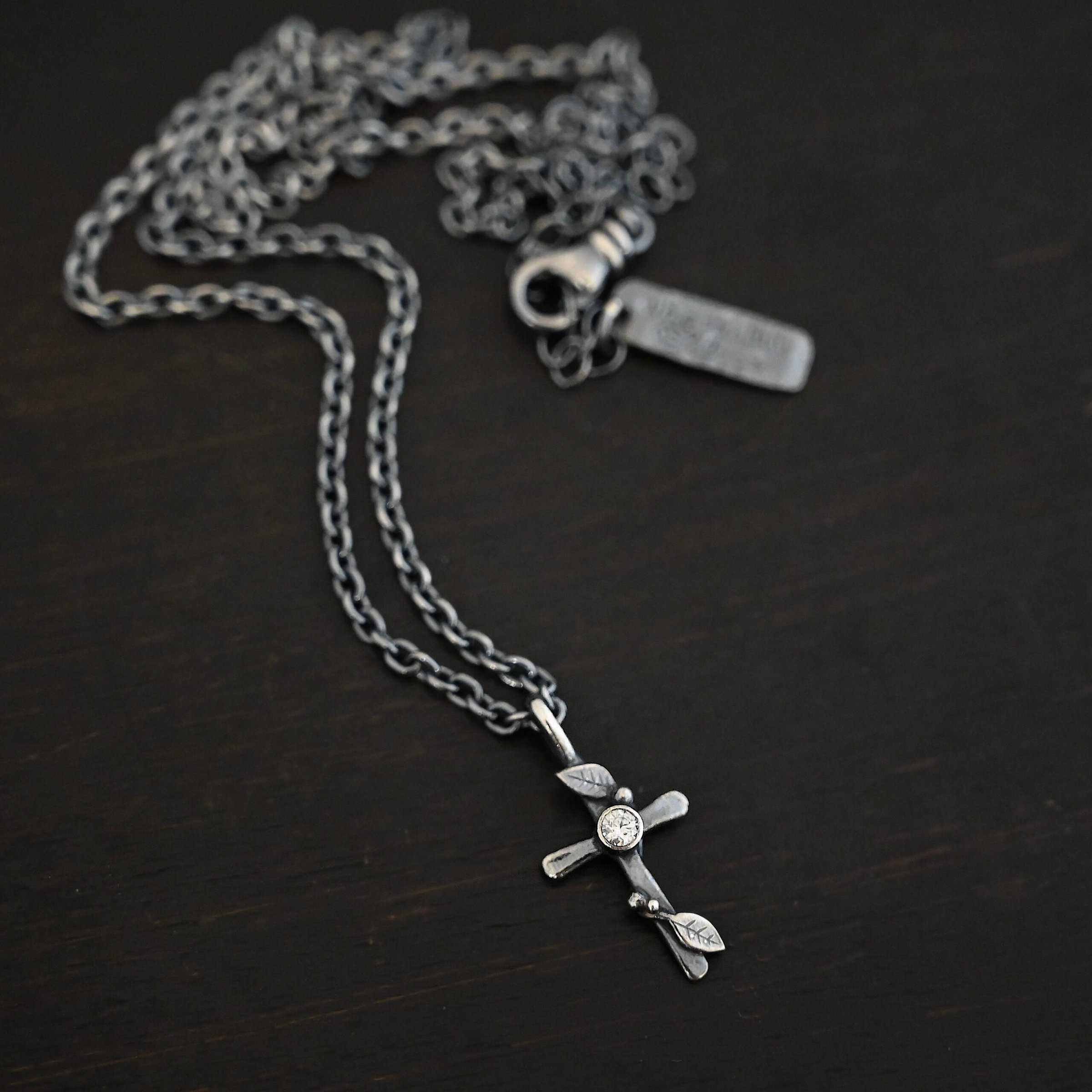 Third Day Necklace Photo of "The Third Day" Necklace. Made out of sterling silver and a moissanite. The pendant is a sterling silver cross with silver vines and leaves wrapped all around it. There is a small diamond in the center of the cross. There is a loop on top of the cross that is attached to a silver jump ring that are attached to the chain of the necklace. Necklace is laying against a black background.