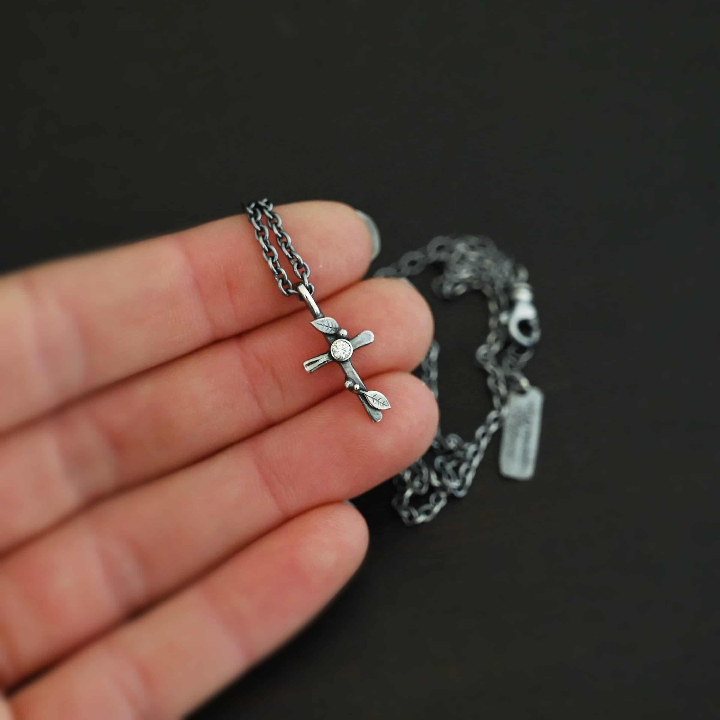Third Day Necklace Photo of "The Third Day" Necklace. Made out of sterling silver and a moissanite. The pendant is a sterling silver cross with silver vines and leaves wrapped all around it. There is a small diamond in the center of the cross. There is a loop on top of the cross that is attached to a silver jump ring that are attached to the chain of the necklace. Necklace is held in the fingertips of a hand to demonstrate size.