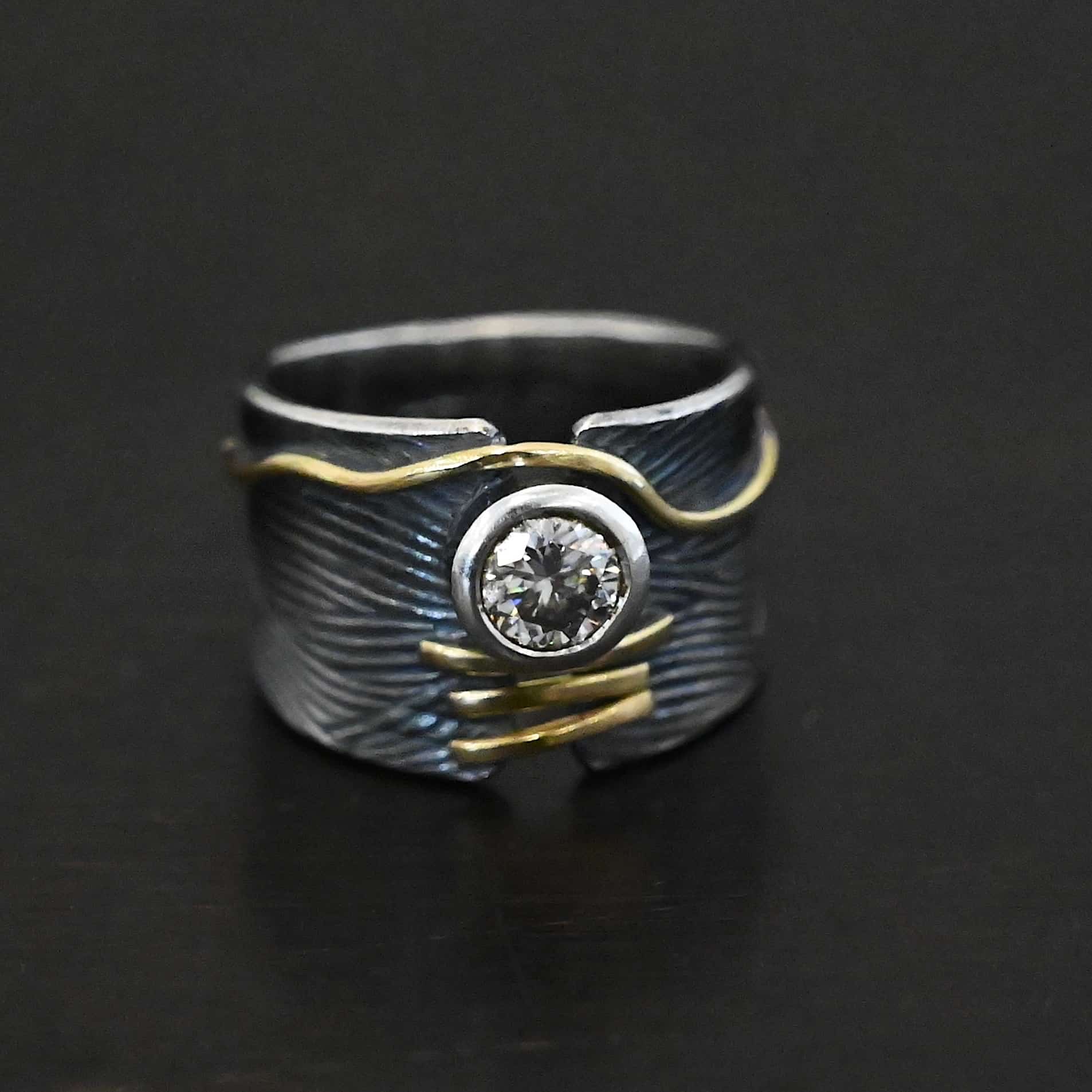 Endless Love Engagement Ring Photo of the "Endless Love" Engagement Ring. Made out of sterling silver, gold, and moissanite. The moissanite is in the center of the ring. There are three lines of gold underneath it, connecting the gap in the ring band together. There is a long line of gold above the moissanite that curves around it and goes around the entire band. This line of gold flows up and down as it goes around the band. The rest of the band is silver and has wavy lines engraved in it as well. The center of the ring band has a gap in which the moissanite and gold bands are connecting it together. Ring sits against a dark background.