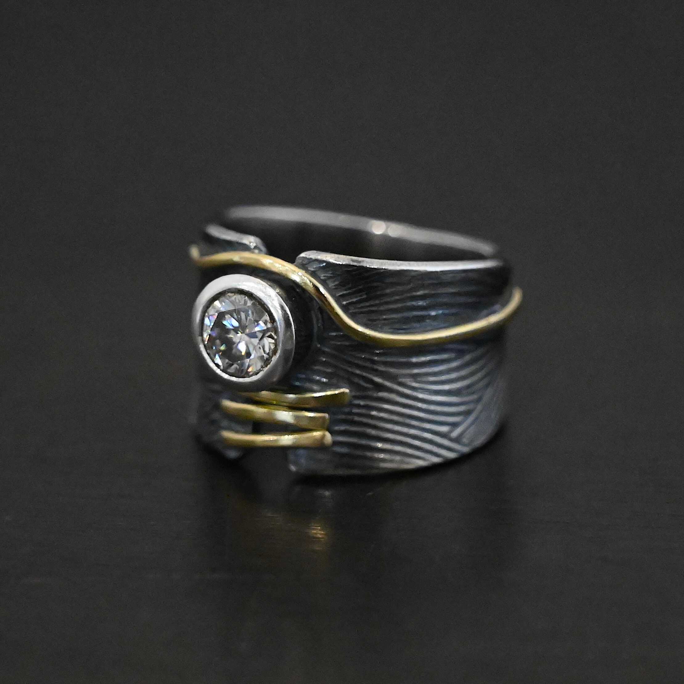 Endless Love Engagement Ring Photo of the "Endless Love" Engagement Ring. Made out of sterling silver, gold, and moissanite. The moissanite is in the center of the ring. There are three lines of gold underneath it, connecting the gap in the ring band together. There is a long line of gold above the moissanite that curves around it and goes around the entire band. This line of gold flows up and down as it goes around the band. The rest of the band is silver and has wavy lines engraved in it as well. The center of the ring band has a gap in which the moissanite and gold bands are connecting it together. Ring sits against a dark background.