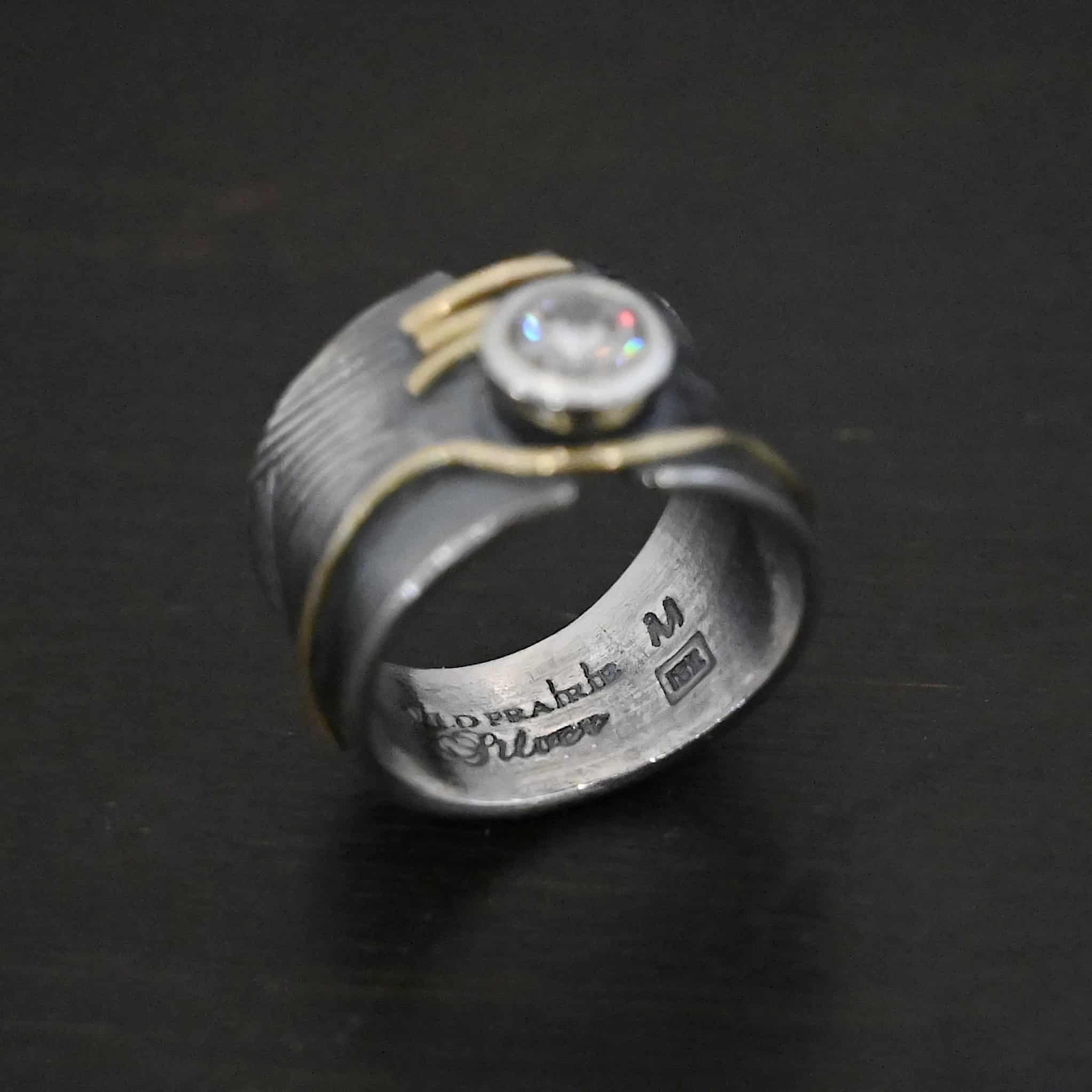 Endless Love Engagement Ring Photo of the "Endless Love" Engagement Ring. Made out of sterling silver, gold, and moissanite. The moissanite is in the center of the ring. There are three lines of gold underneath it, connecting the gap in the ring band together. There is a long line of gold above the moissanite that curves around it and goes around the entire band. This line of gold flows up and down as it goes around the band. The rest of the band is silver and has wavy lines engraved in it as well. The center of the ring band has a gap in which the moissanite and gold bands are connecting it together. Ring sits against a dark background. The words "Wild Prairie Silver" are engraved on the inside, as well as the letter "M" and a rectangle box with another "M" in that.
