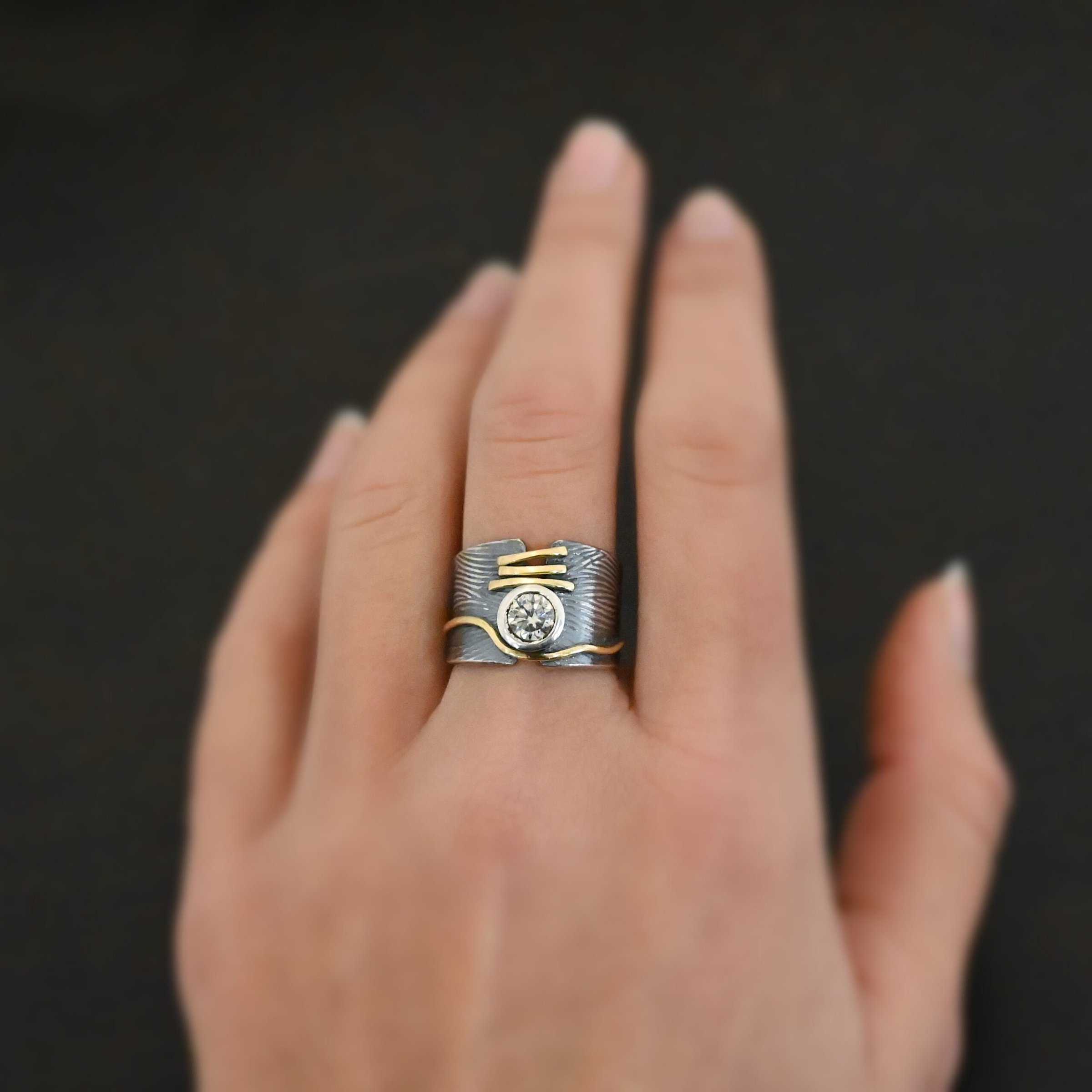 Endless Love Engagement Ring Photo of the "Endless Love" Engagement Ring. Made out of sterling silver, gold, and moissanite. The moissanite is in the center of the ring. There are three lines of gold underneath it, connecting the gap in the ring band together. There is a long line of gold above the moissanite that curves around it and goes around the entire band. This line of gold flows up and down as it goes around the band. The rest of the band is silver and has wavy lines engraved in it as well. The center of the ring band has a gap in which the moissanite and gold bands are connecting it together. Ring is worn on a models middle finger to demonstrate size.