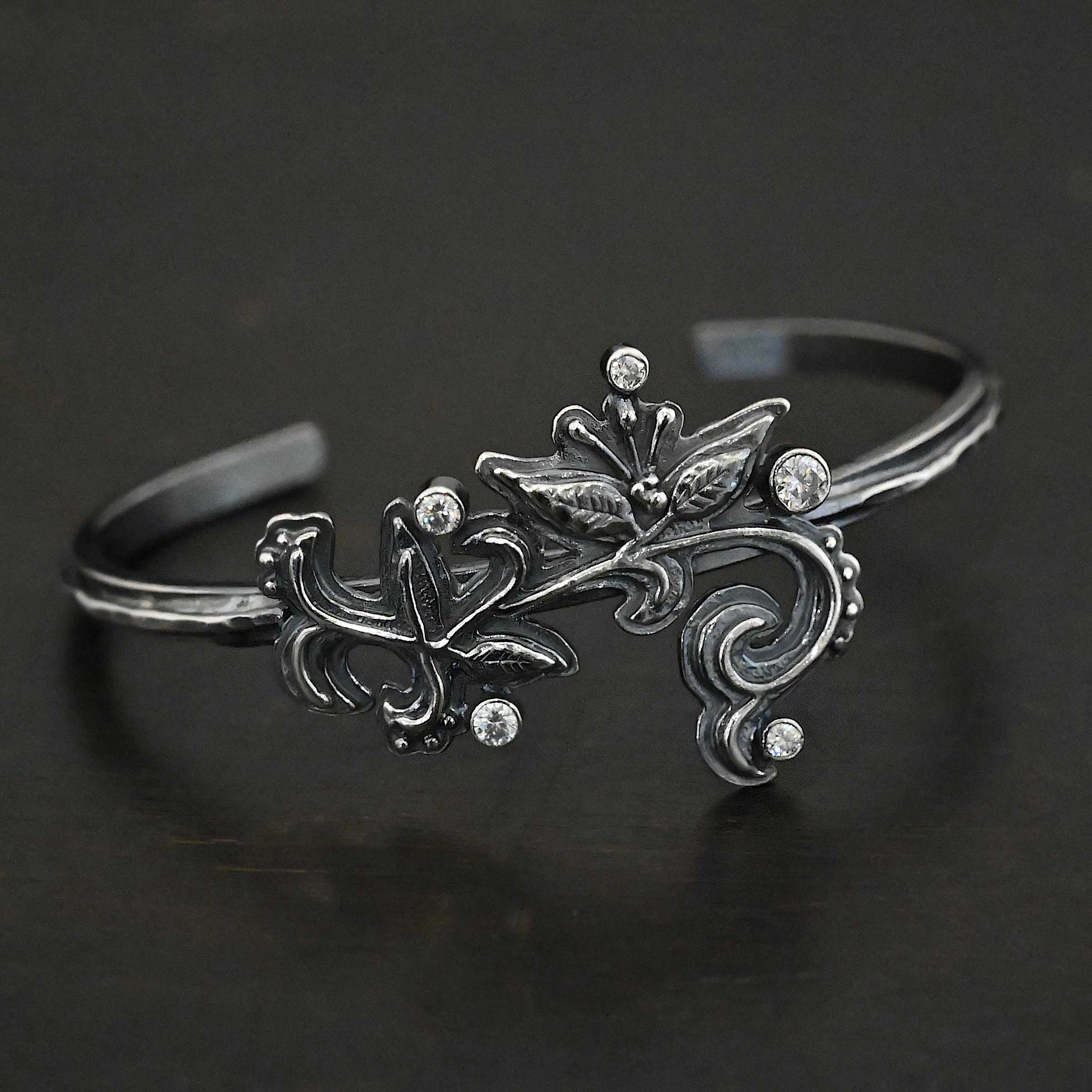 White Satin Cuff Photo of the "White Lace" Cuff. Made out of sterling silver and moissanites. There are five moissanites. They are scattered around a cuff on sterling silver flower and vine like designs that are sawed out and soldered to the cuff. Cuff is placed against a black background.