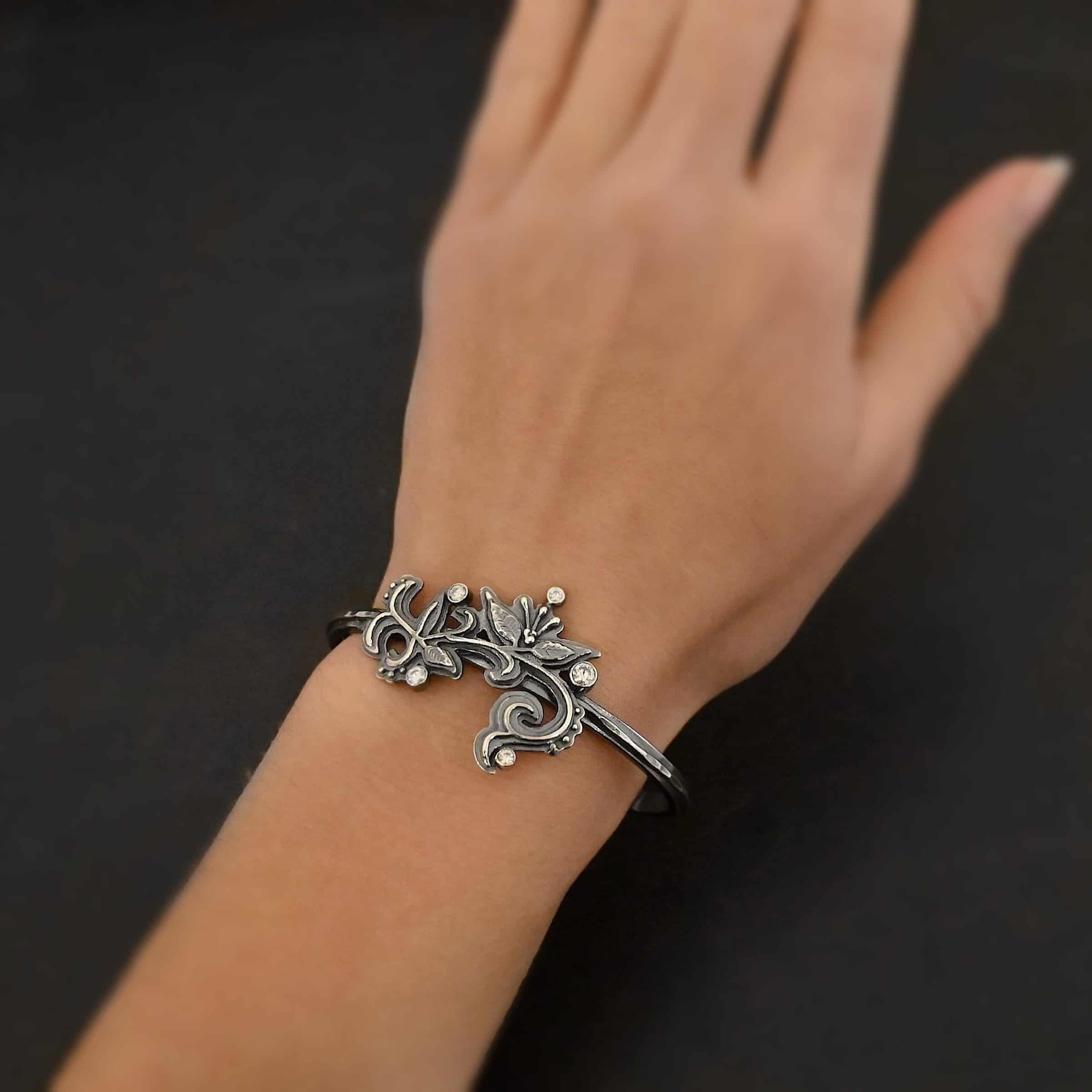 White Satin Cuff Photo of the "White Lace" Cuff. Made out of sterling silver and moissanites. There are five moissanites. They are scattered around a cuff on sterling silver flower and vine like designs that are sawed out and soldered to the cuff. Cuff is worn on a models wrist to demonstrate size.