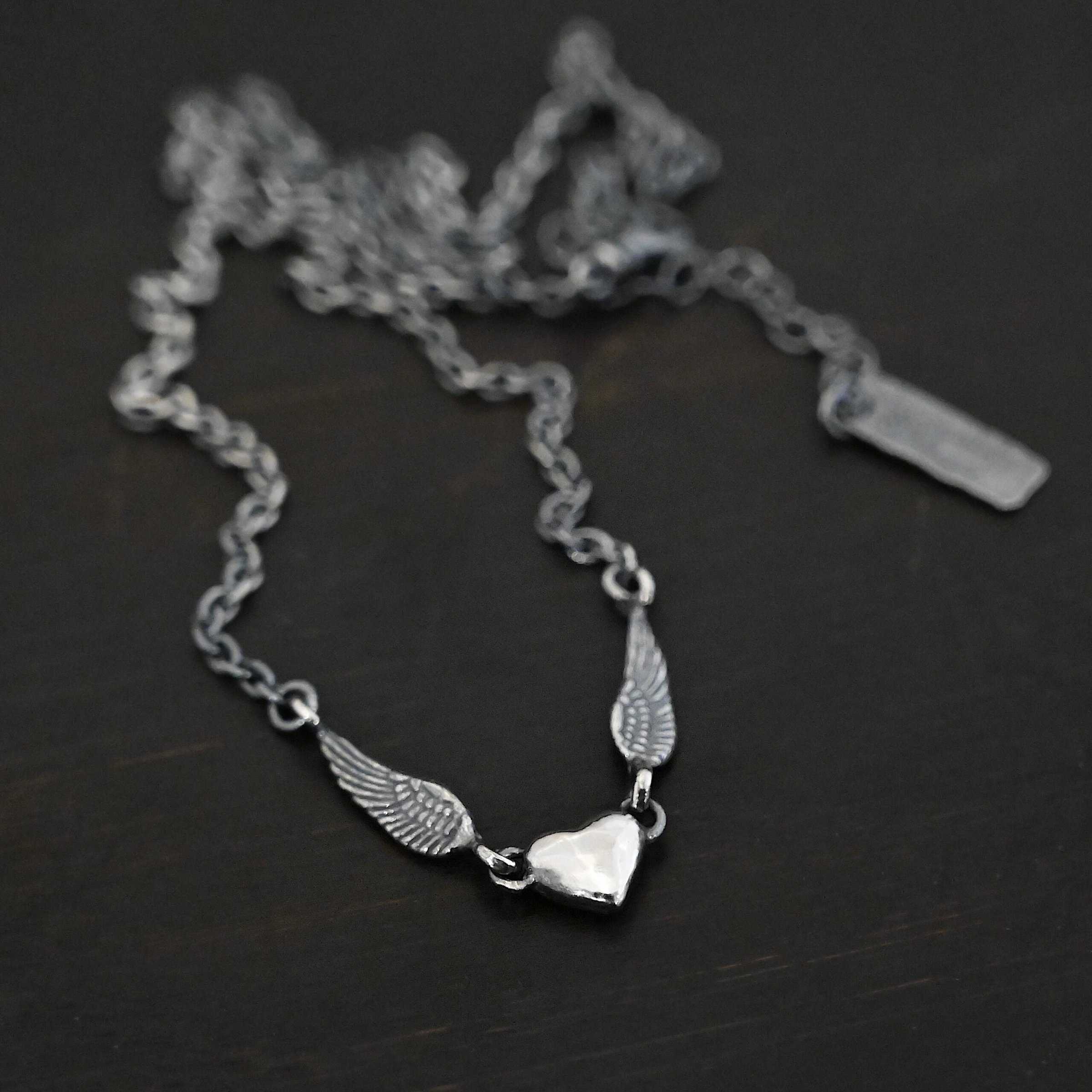 "A Little Courage" Necklace Photo of "A Little Courage" Necklace. Made out of oxidized sterling silver. There is a small heart pendant in the center of the necklace with a loop on each side of it. Attached to each loop is an angel wing. On the end of each angel wing is a loop that the chain of the necklace is attached to. Necklace is laying against a dark background.