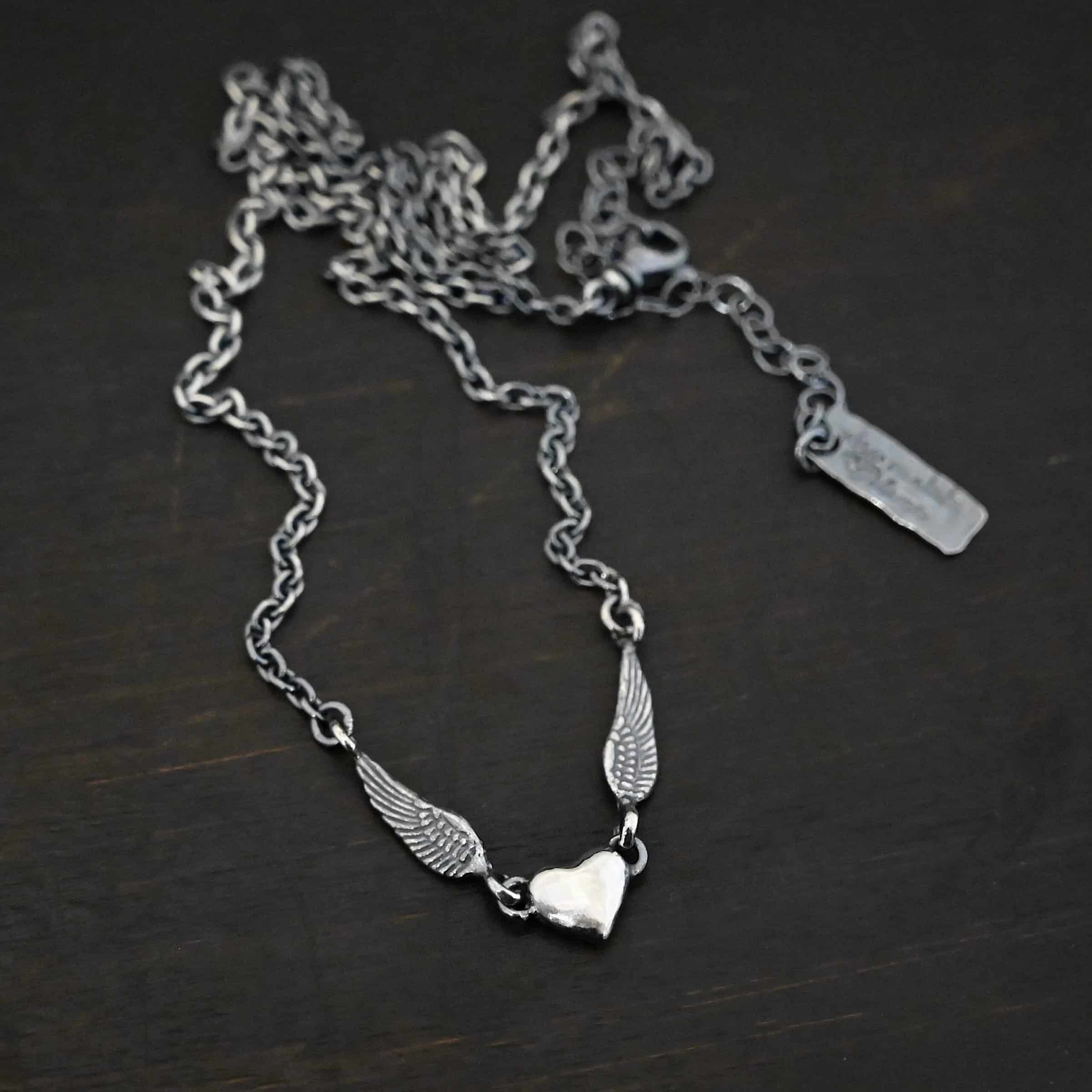 "A Little Courage" Necklace Photo of "A Little Courage" Necklace. Made out of oxidized sterling silver. There is a small heart pendant in the center of the necklace with a loop on each side of it. Attached to each loop is an angel wing. On the end of each angel wing is a loop that the chain of the necklace is attached to. Necklace is laying against a dark background.