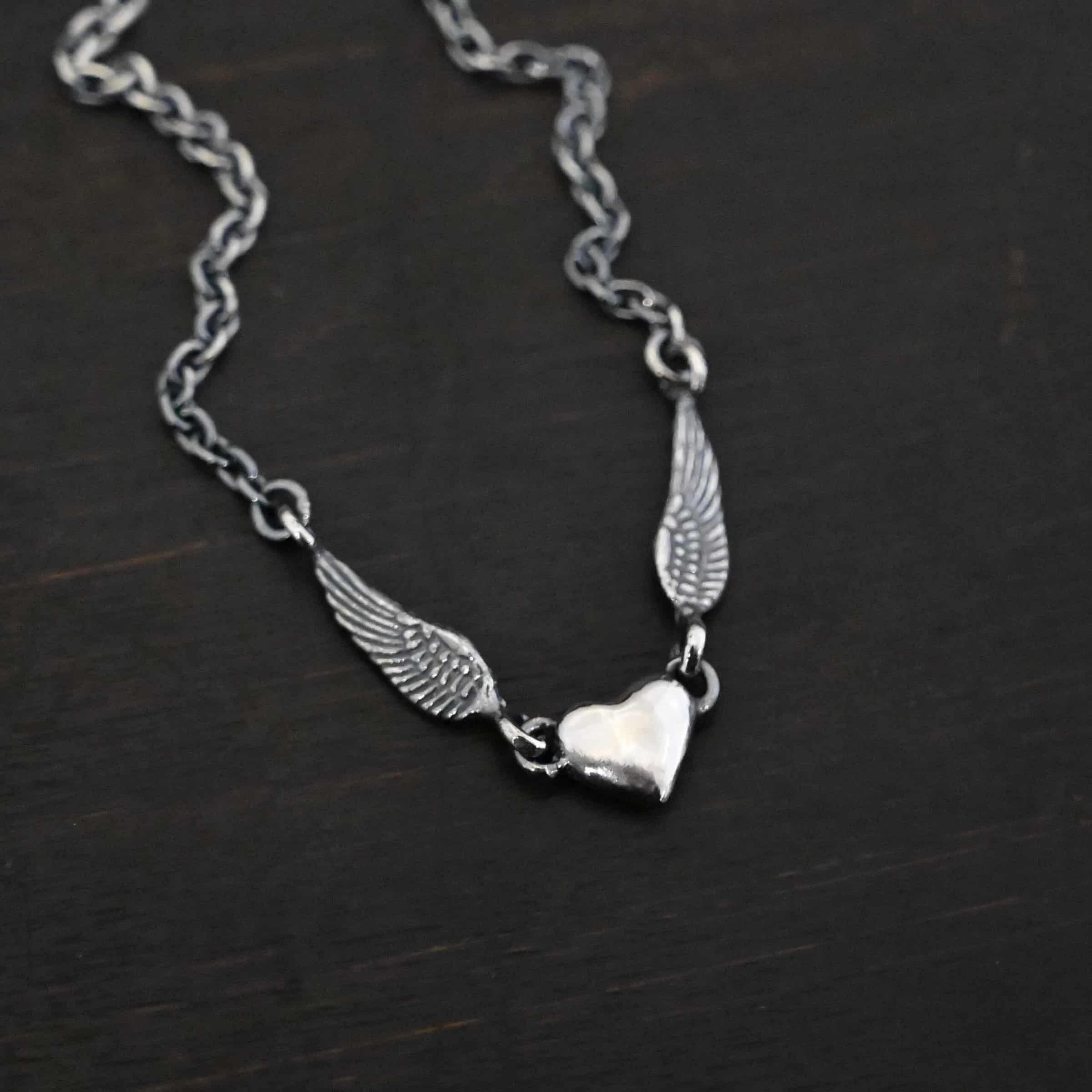 "A Little Courage" Necklace Photo of "A Little Courage" Necklace. Made out of oxidized sterling silver. There is a small heart pendant in the center of the necklace with a loop on each side of it. Attached to each loop is an angel wing. On the end of each angel wing is a loop that the chain of the necklace is attached to. Necklace is laying against a dark background.