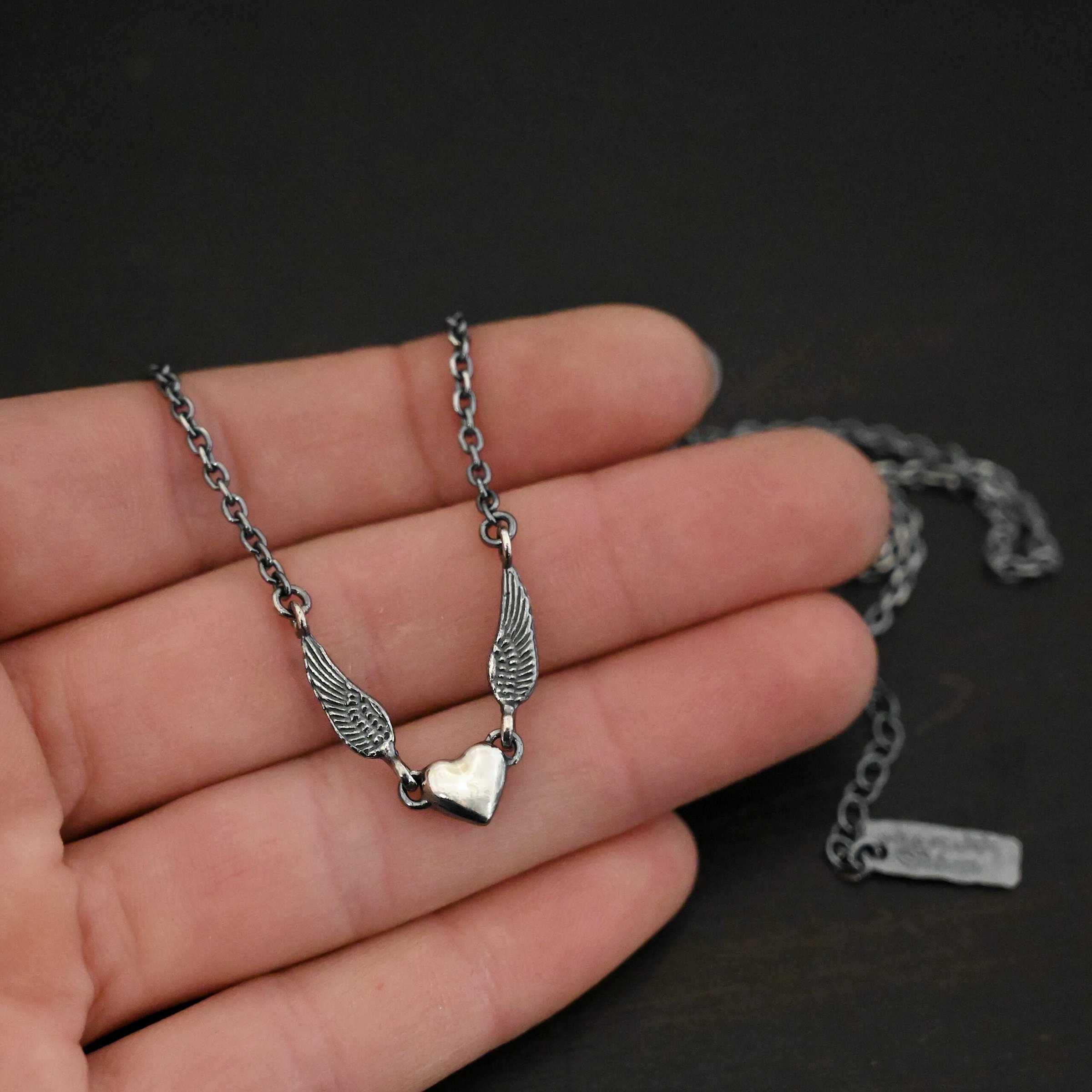 "A Little Courage" Necklace Photo of "A Little Courage" Necklace. Made out of oxidized sterling silver. There is a small heart pendant in the center of the necklace with a loop on each side of it. Attached to each loop is an angel wing. On the end of each angel wing is a loop that the chain of the necklace is attached to. Necklace is laying against the fingertips of a hand.