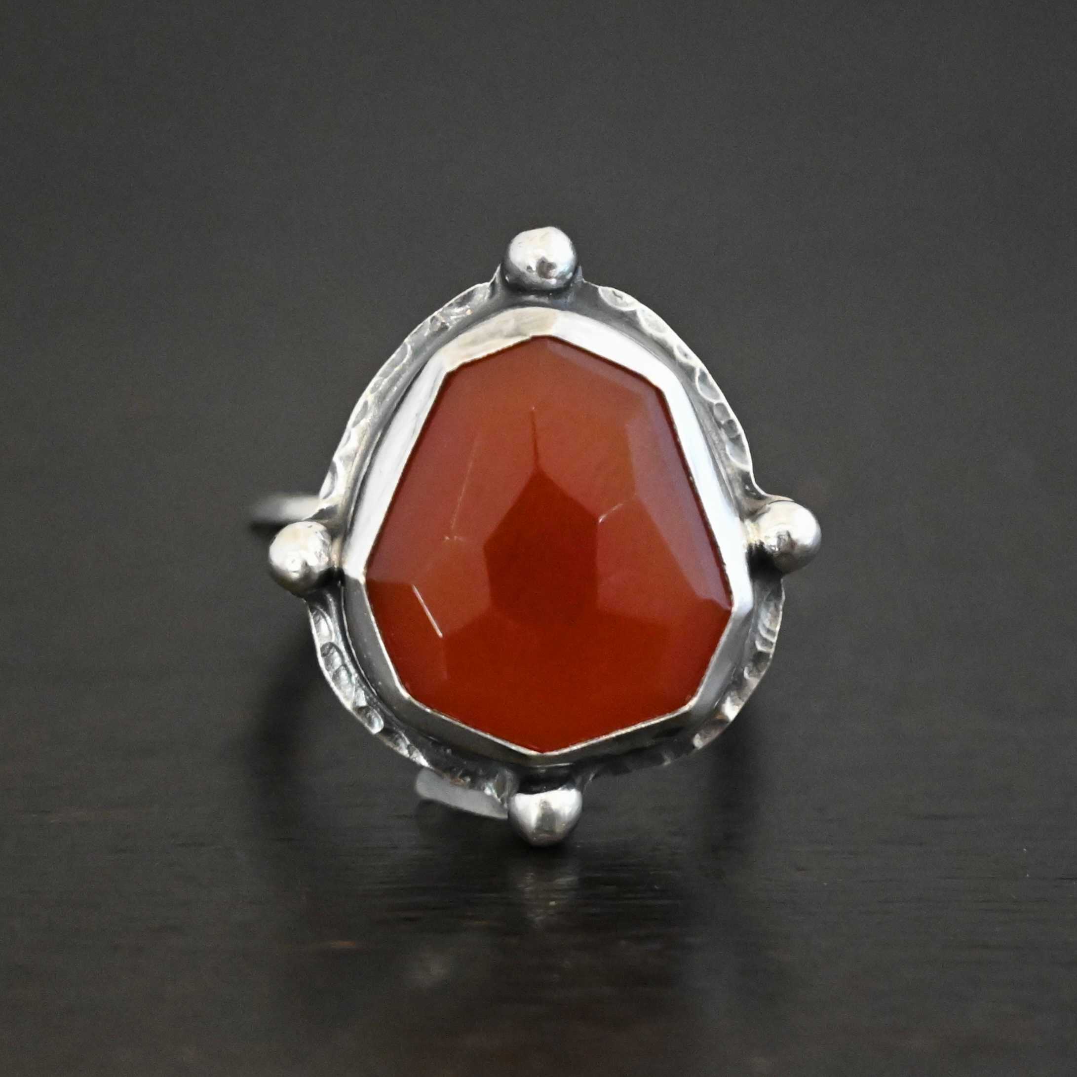 Cindy Lou Ring Photo of the "Cindy Lou" Ring. Made out of sterling silver and carnelian. The carnelian is a deep orange color and rose cut. The silver setting around it is hammered. On each side of the carnelian is a silver dot. One on the top, one on each side, and one on the bottom. Ring has a simple silver band. Ring is sitting against a dark background.