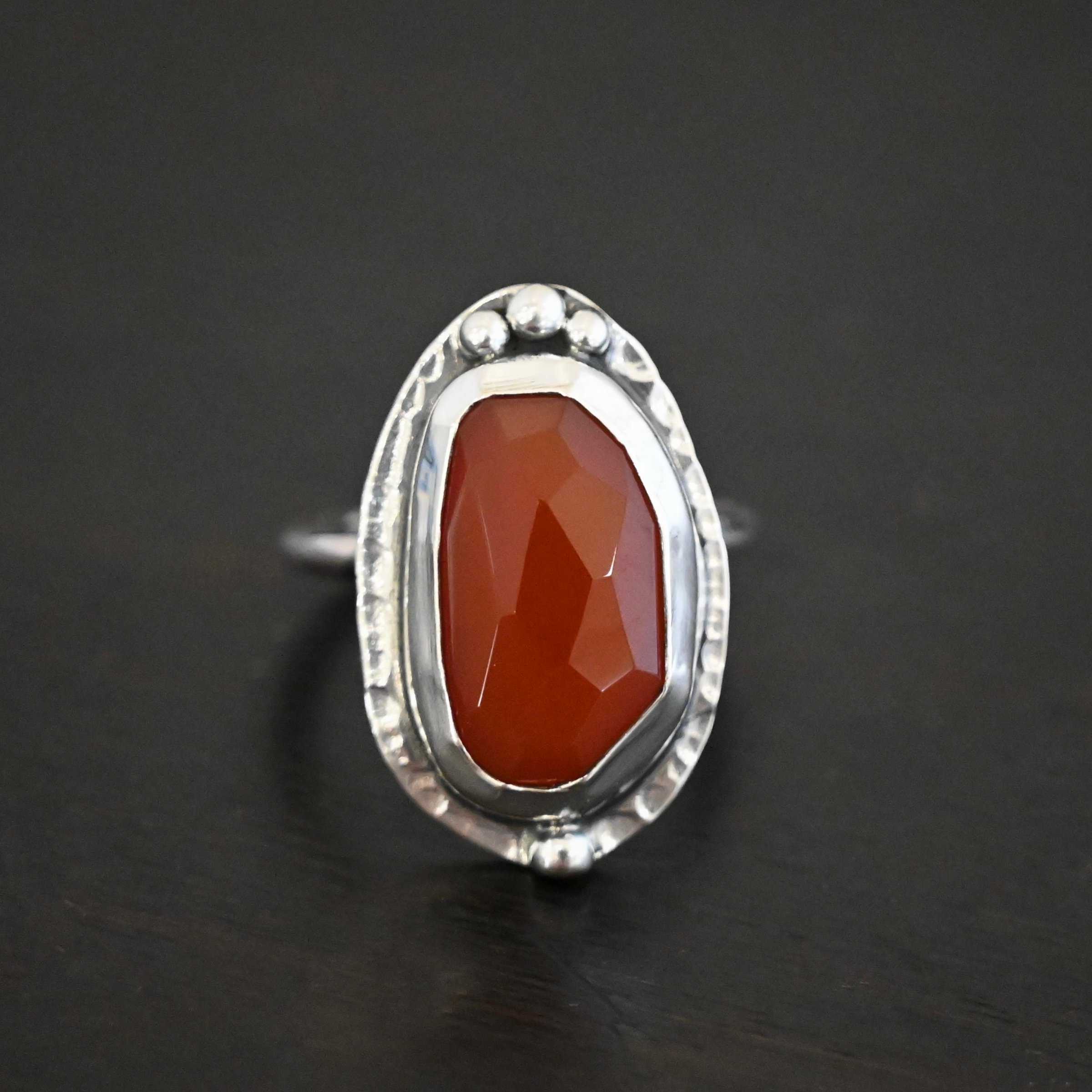 Clarice Ring Photo of the "Clarice" Ring. Made out of sterling silver and carnelian. The carnelian is a deep orange color and an oval shape. It is also rose cut. The silver setting around it is hammered. On the top of the carnelian, following the curve of it are three silver dots set right next to one another. Ring has a simple silver band. Ring is sitting against a dark background.