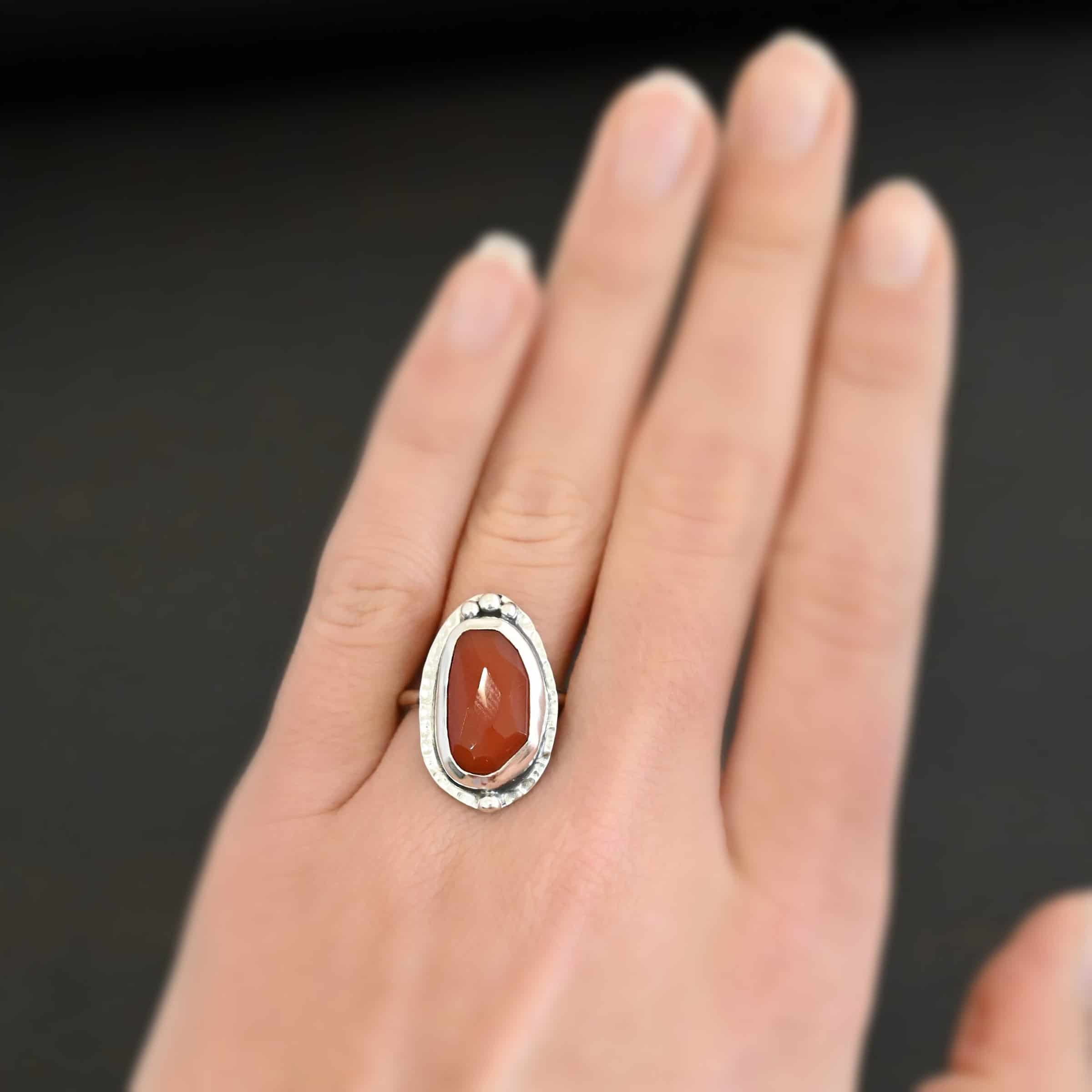 Clarice Ring Photo of the "Clarice" Ring. Made out of sterling silver and carnelian. The carnelian is a deep orange color and an oval shape. It is also rose cut. The silver setting around it is hammered. On the top of the carnelian, following the curve of it are three silver dots set right next to one another. Ring has a simple silver band. Ring is worn on a models middle finger to demonstrate size.