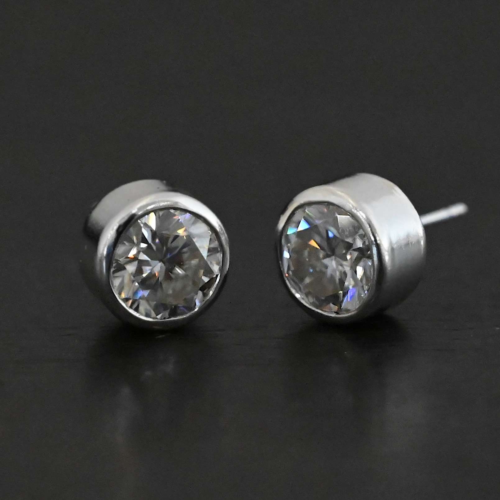 Photo of the "Betina" Earrings. Made out of sterling silver and moissanites. Earrings are sterling silver post earrings with moissanites in the center of them. The settings for the moissanites are thick and the posts are on the back center of the setting. Earrings are laying against a dark background.