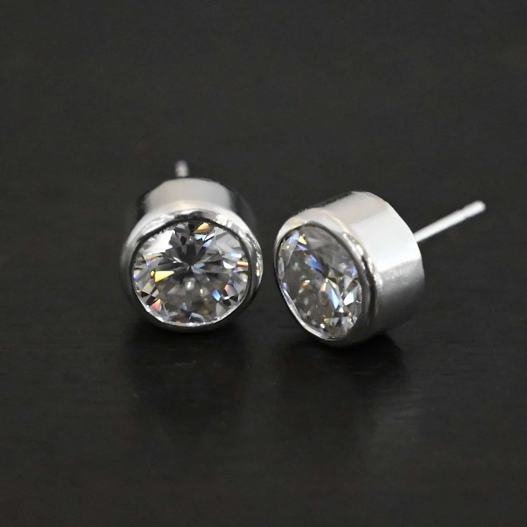 Photo of the "Betina" Earrings. Made out of sterling silver and moissanites. Earrings are sterling silver post earrings with moissanites in the center of them. The settings for the moissanites are thick and the posts are on the back center of the setting. Earrings are laying against a dark background.